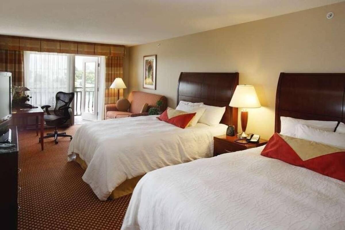 A hotel room with two beds, a desk, a chair, a lamp, and a window.