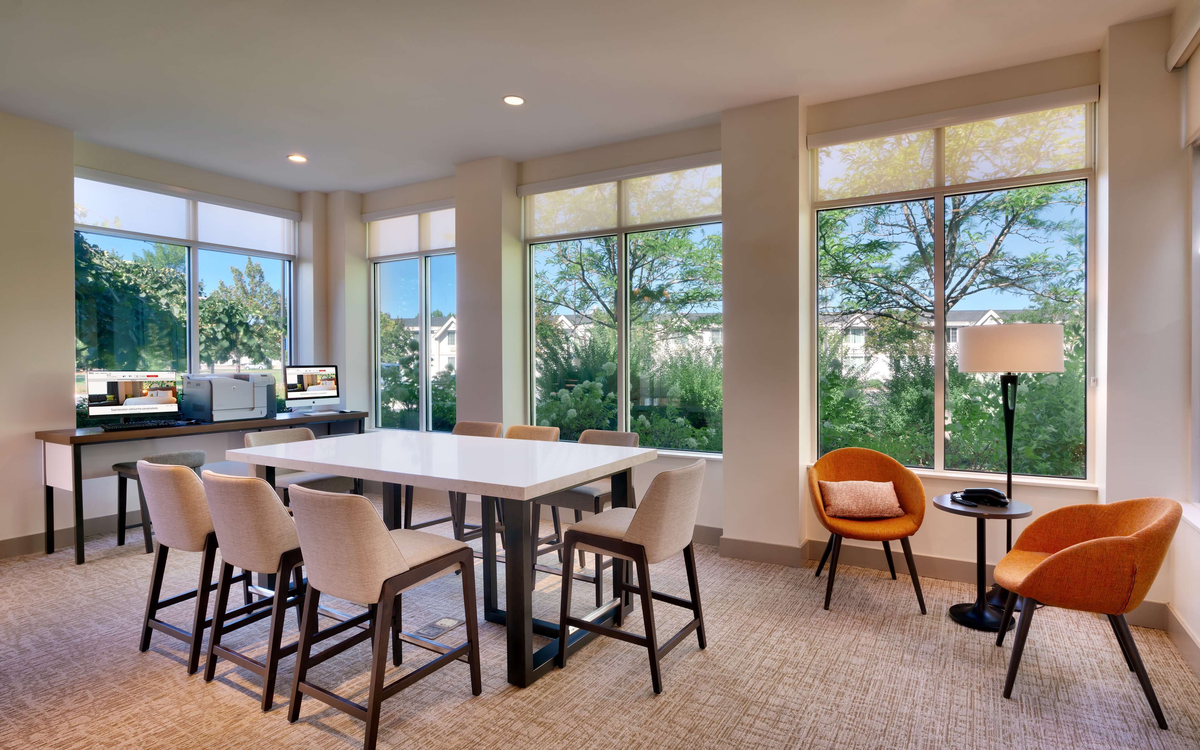 A spacious room with a large table and chairs, a desk with a computer, and large windows offering a view of trees and buildings.
