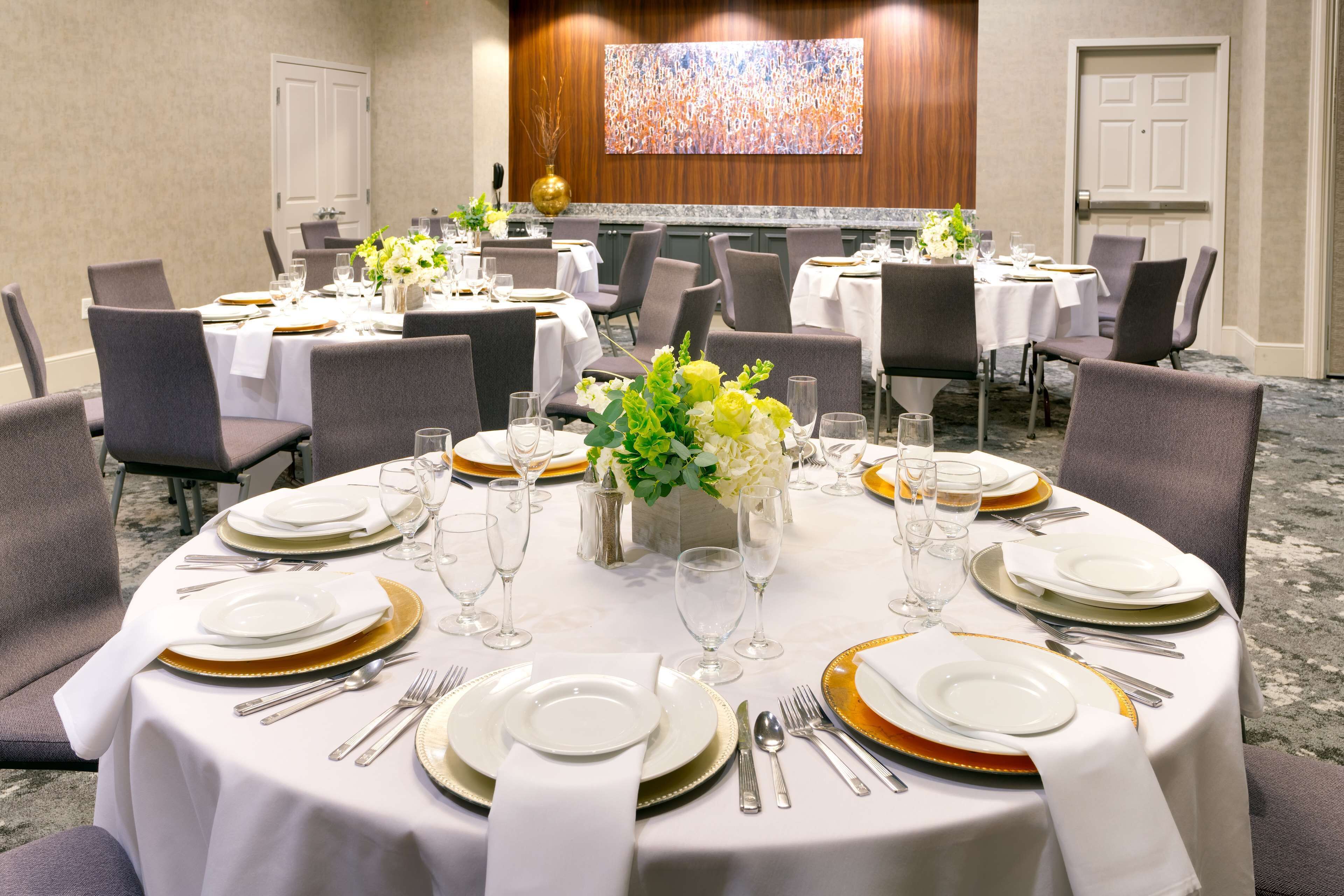 Un salón de conferencias con mesas redondas preparadas para un evento formal, con manteles blancos, platos con borde dorado y cubertería.