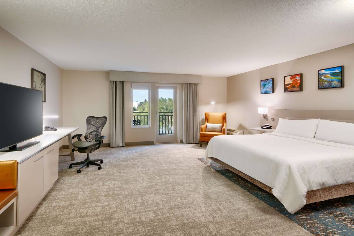 A hotel room with a bed, a desk with a TV, a chair, a balcony with curtains, and artwork on the walls.
