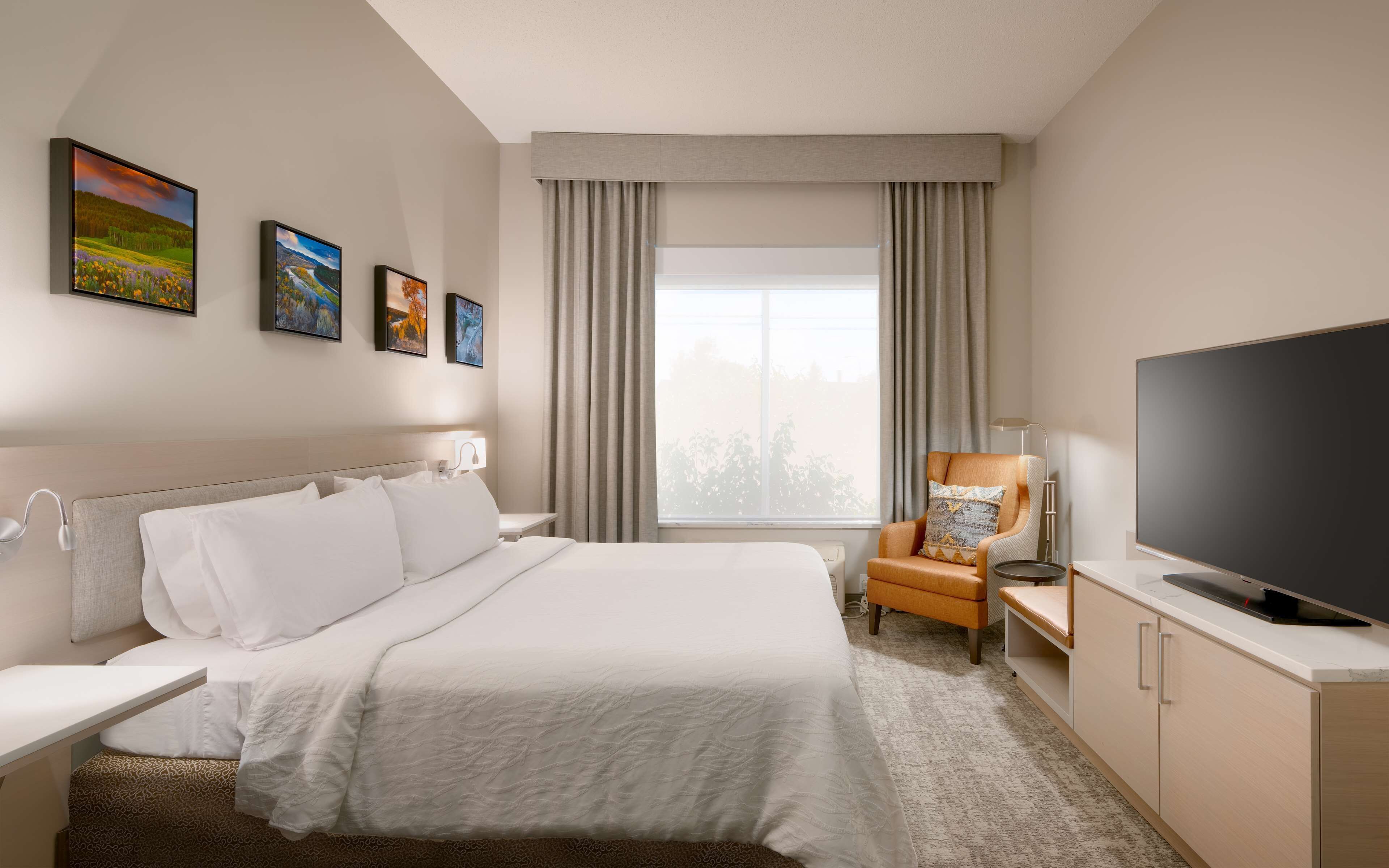 A hotel room with a bed, a desk with a chair, a television, and three paintings on the wall.
