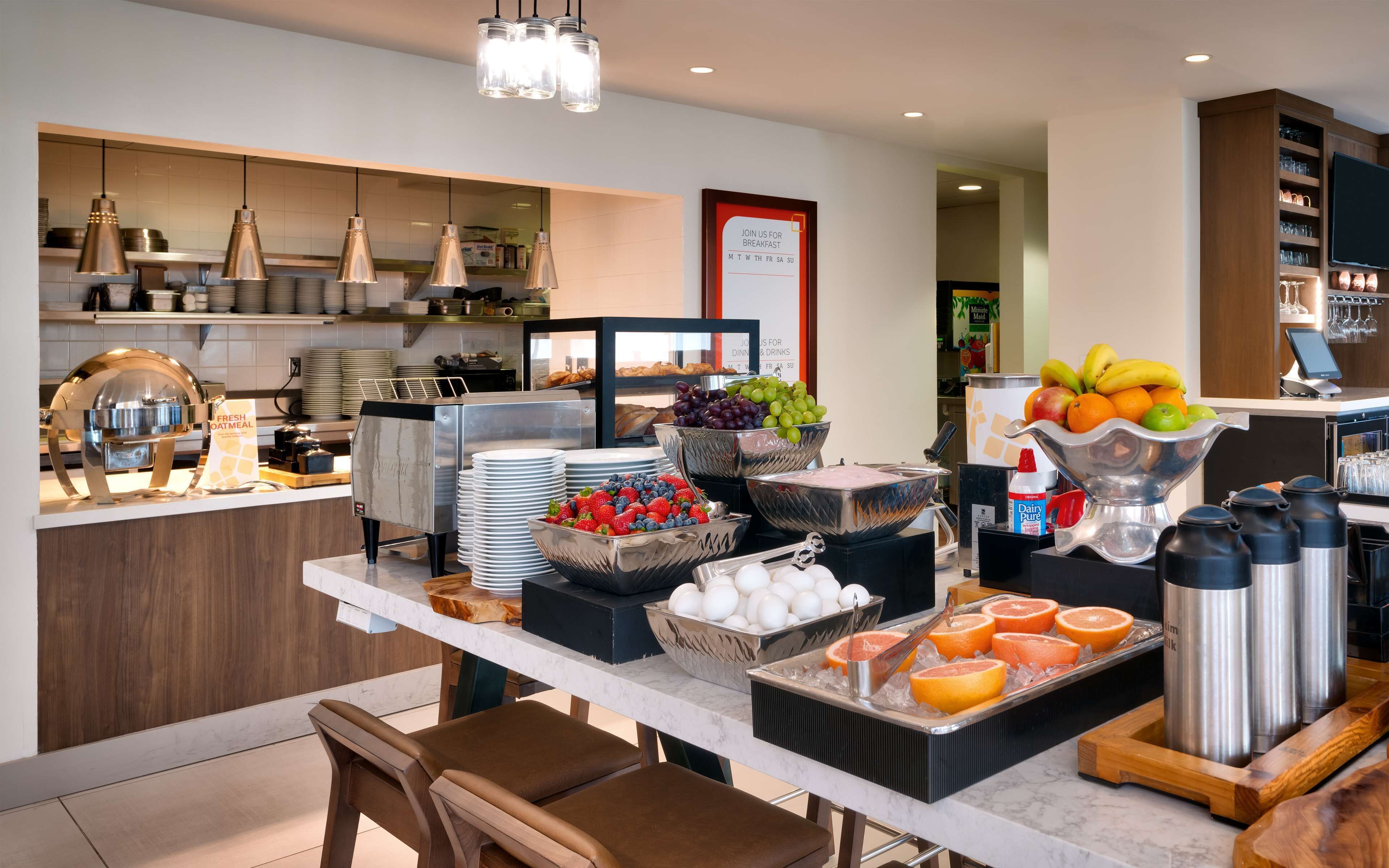 Un montaje tipo bufé con diversos alimentos, entre ellos frutas, huevos y productos lácteos, y un letrero que indica los horarios del desayuno.