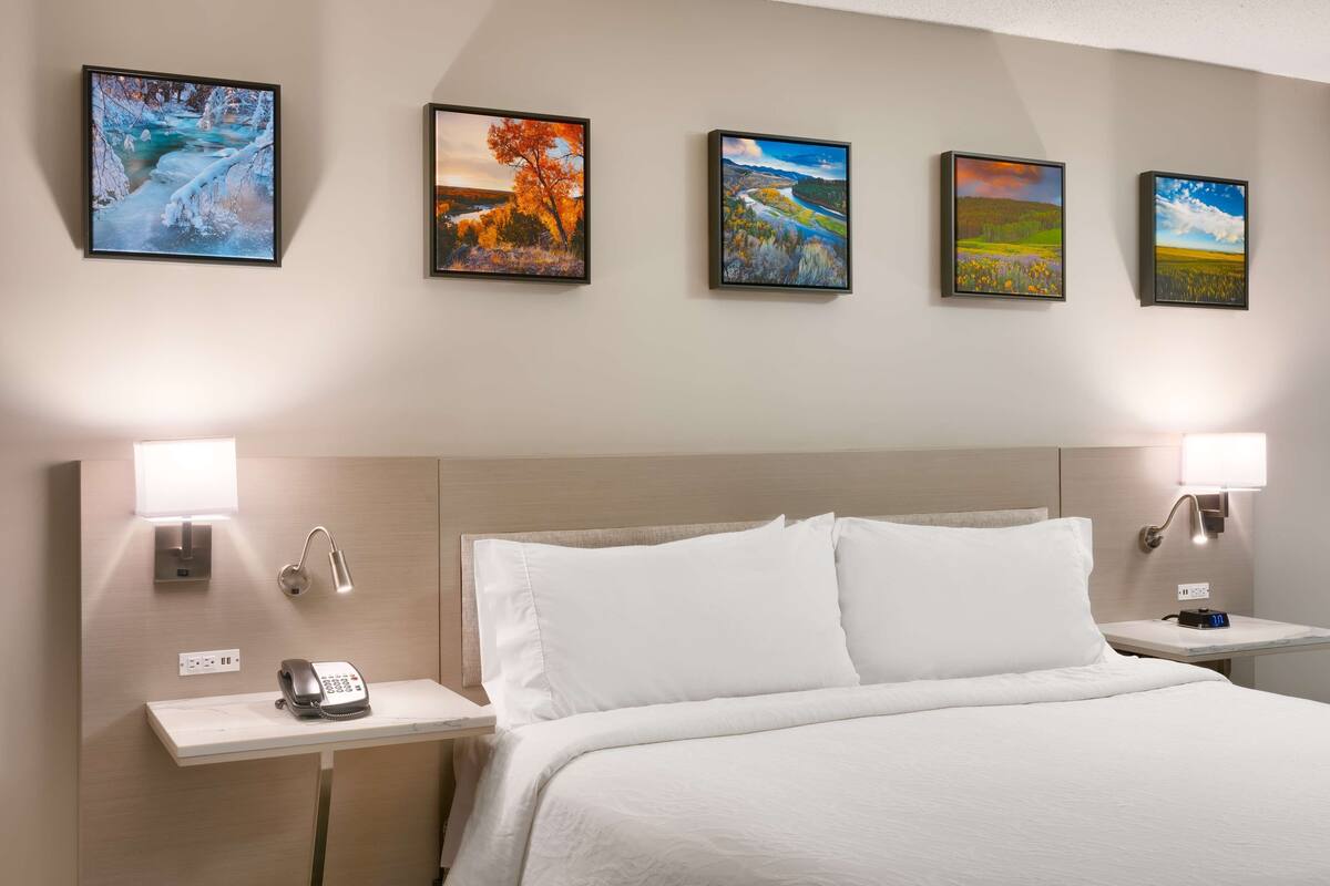 A hotel room with a large bed, two bedside tables with lamps, a telephone, and four landscape paintings on the wall.