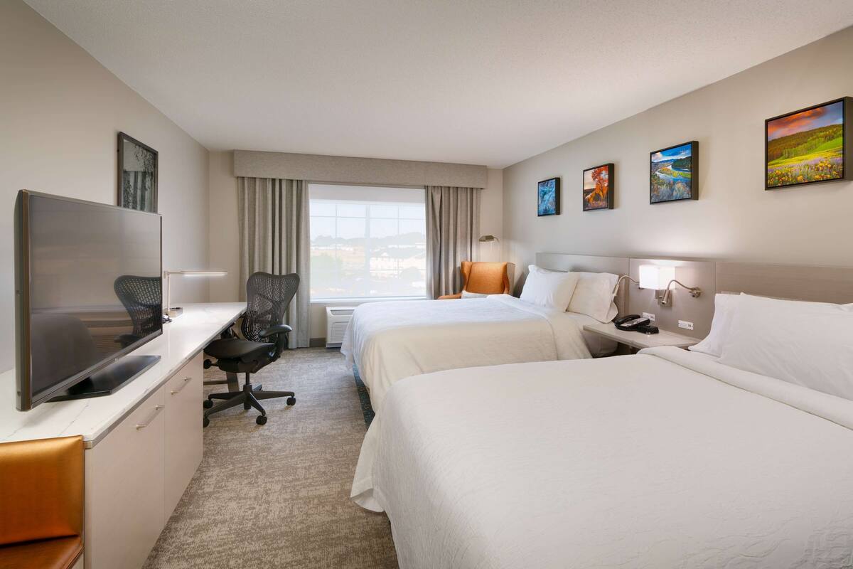 A hotel room with two beds, a desk with a flat-screen TV, a chair, a bedside table with a lamp, and artwork on the walls.
