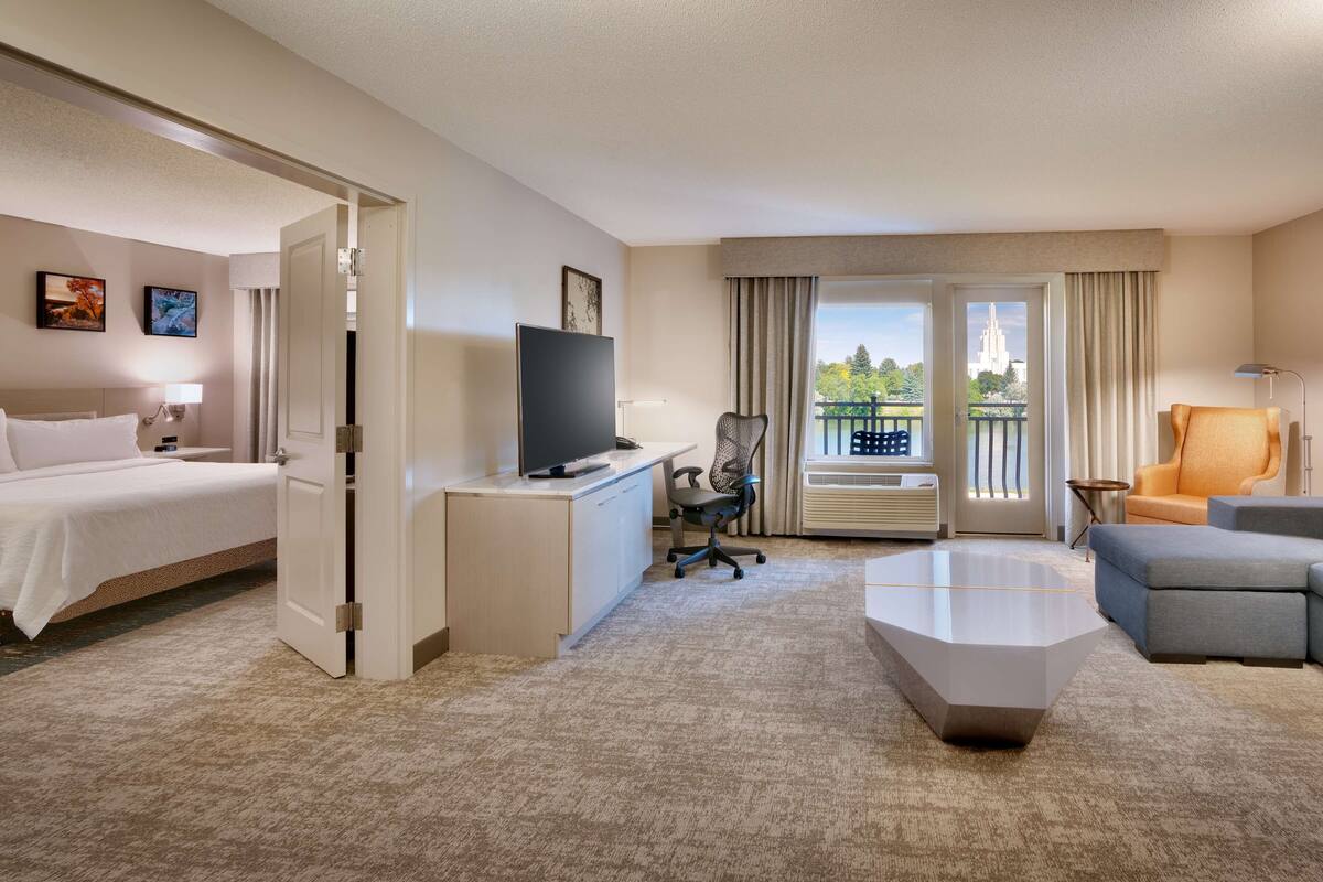 A hotel room with a bed, desk, TV, office chair, sofa, and a view of a balcony with outdoor furniture.