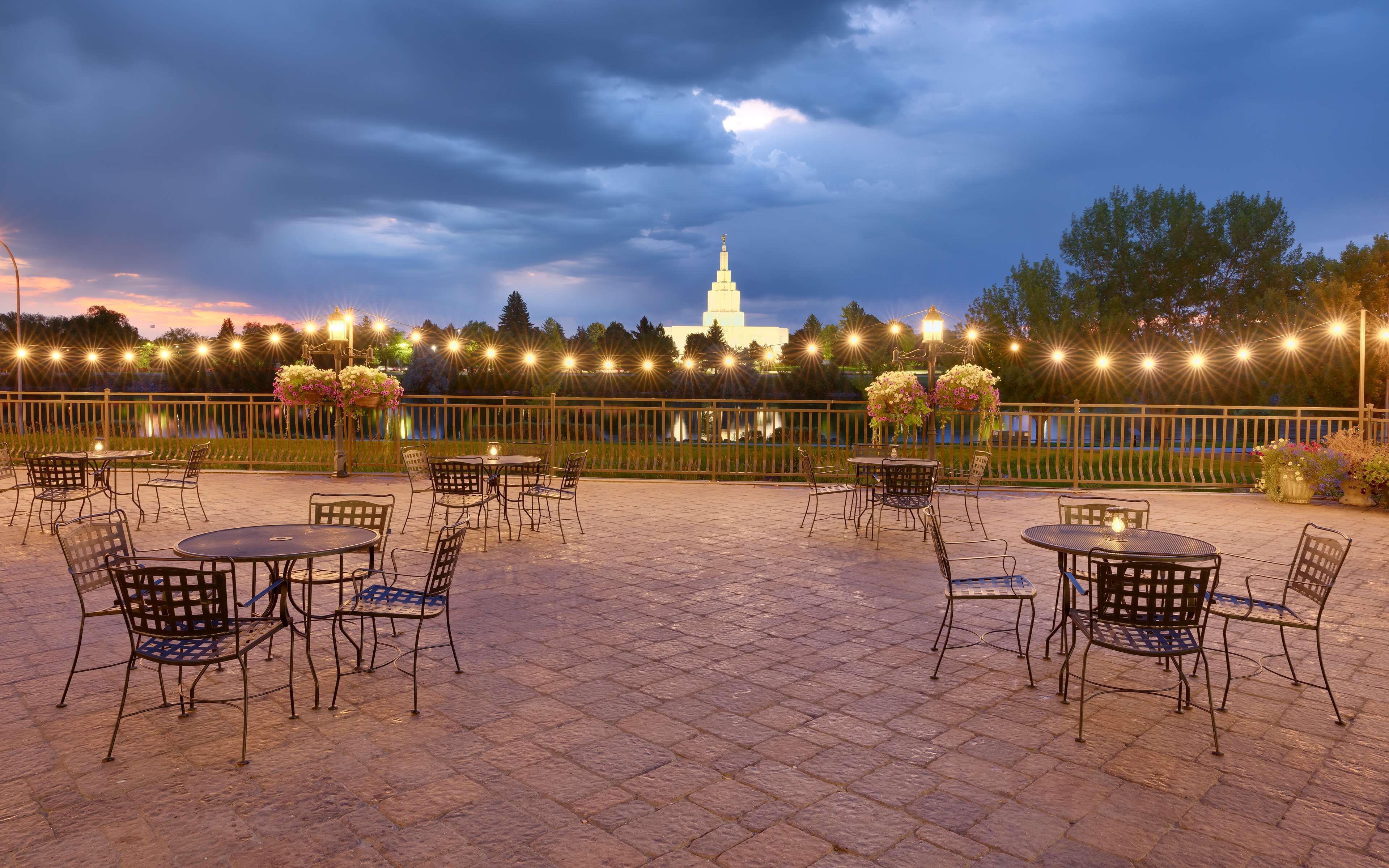 Un patio al aire libre con mesas y sillas, vista a un edificio blanco y luces colgantes al fondo.