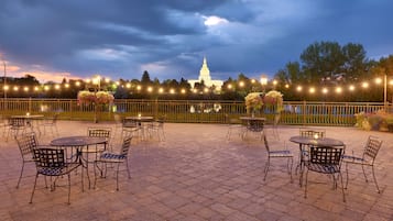 Un patio al aire libre con mesas y sillas, vista a un edificio blanco y luces colgantes al fondo.