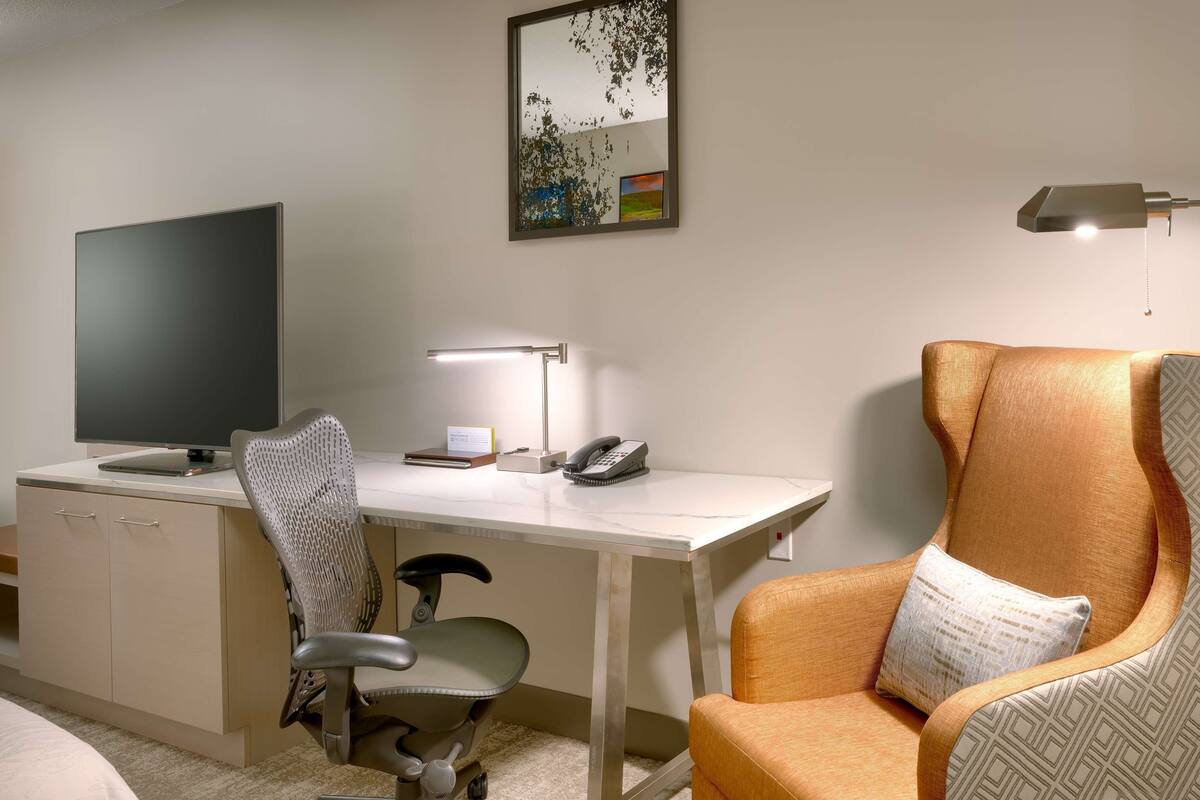 A hotel room with a desk, a computer, a telephone, a lamp, and two chairs.