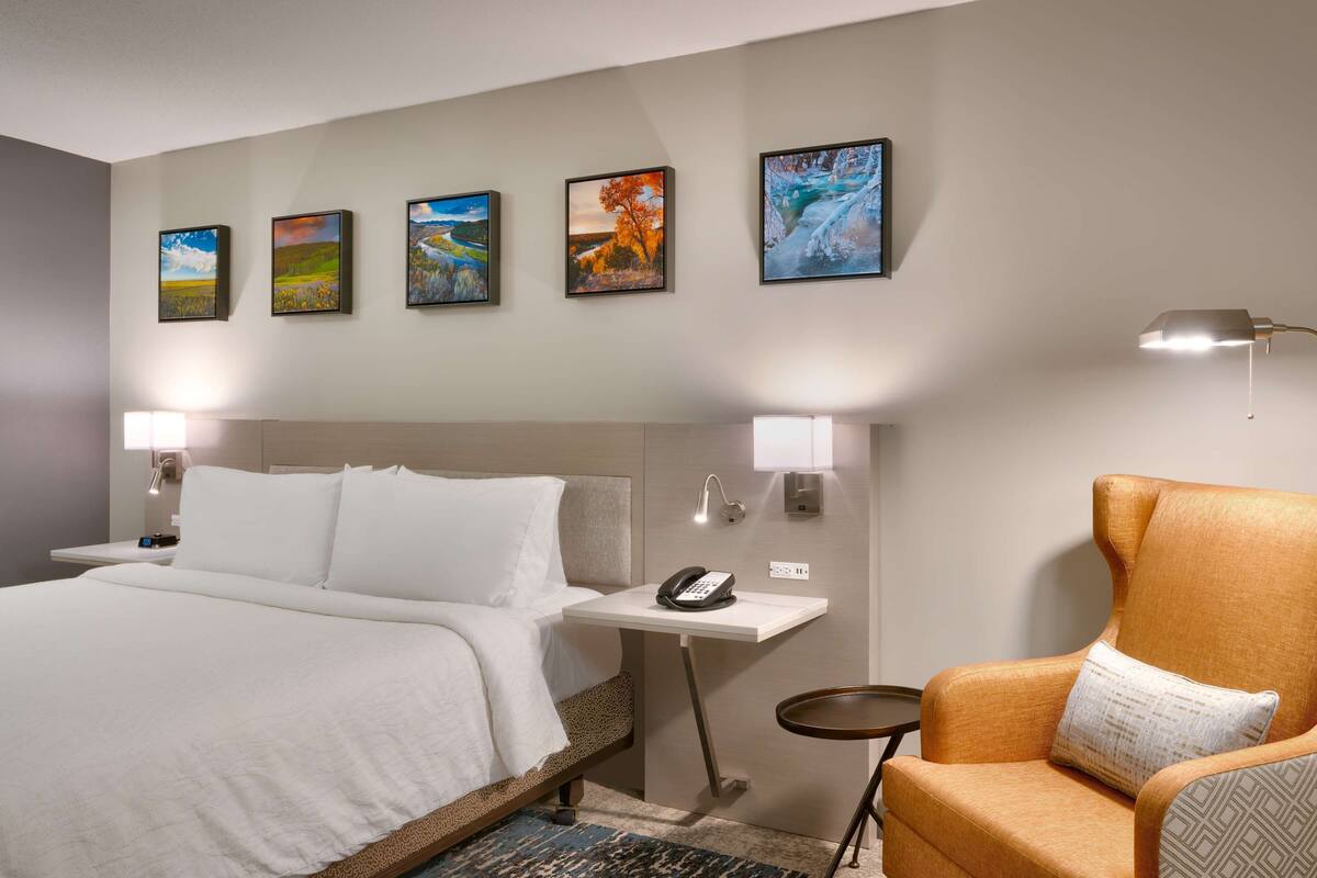 A hotel room with a bed, a chair, a desk with a phone, and wall-mounted lamps.