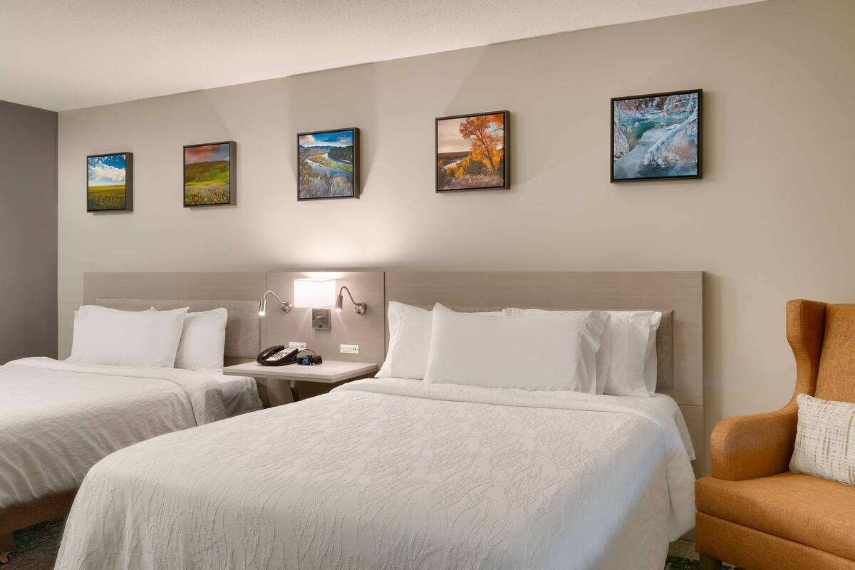 A hotel room with two beds, a nightstand, a lamp, and a chair.