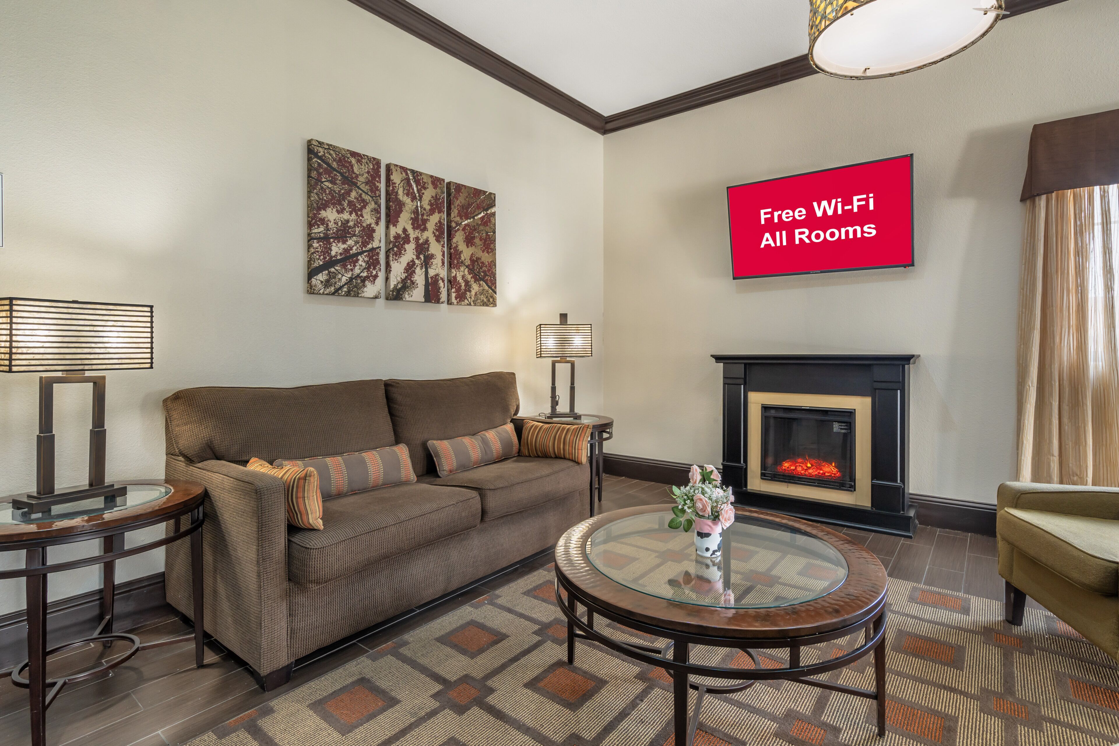 A living room with a brown sofa, a glass coffee table, a fireplace, and a flat-screen TV displaying 'Free Wi-Fi All Rooms'.
