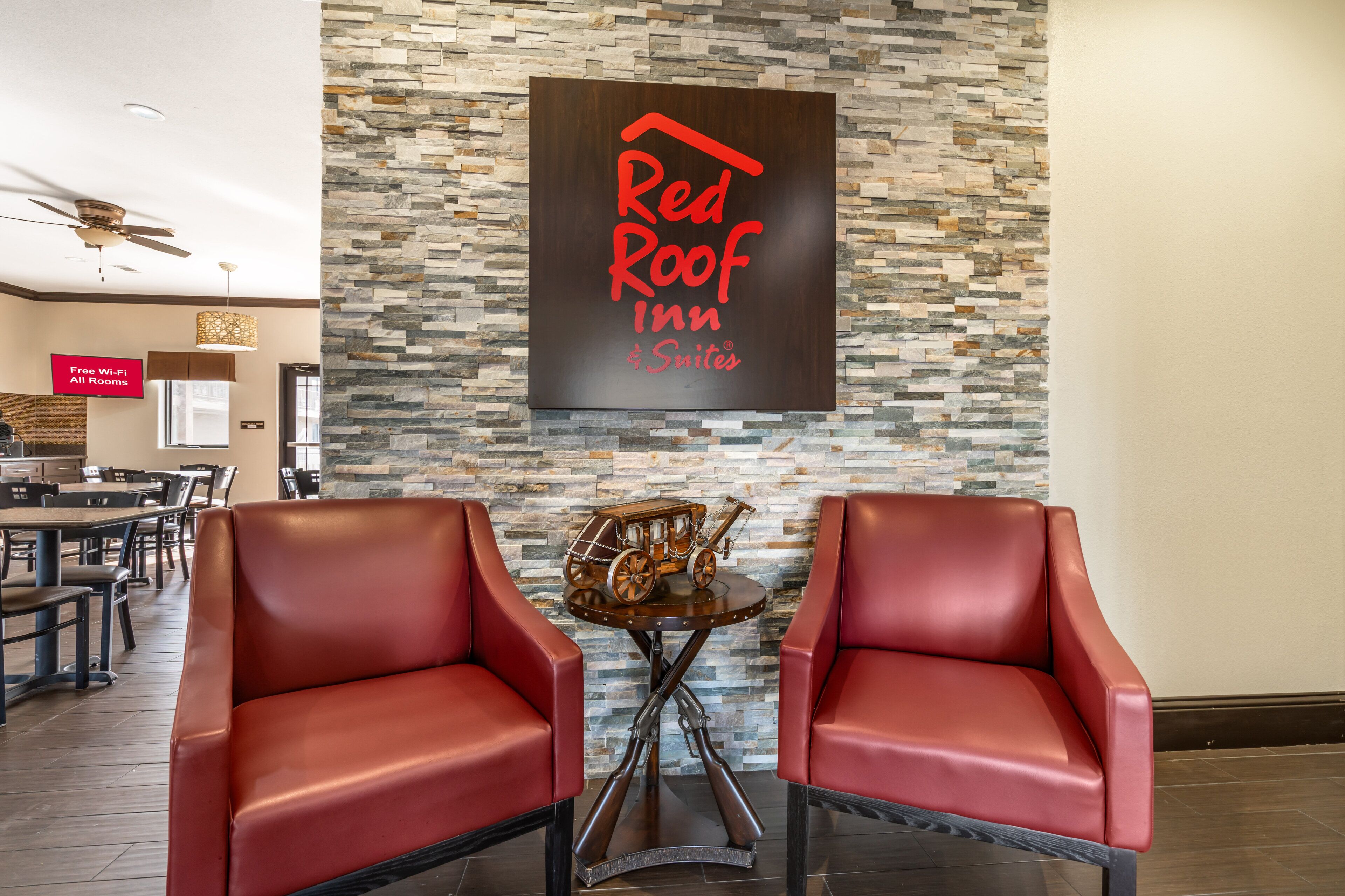 Letrero de Red Roof Inn & Suites sobre una pared de piedra, dos sillones de cuero, una mesita con un modelo antiguo de carruaje, y un letrero que dice 'Wi-Fi Gratis en Todas las Habitaciones'.