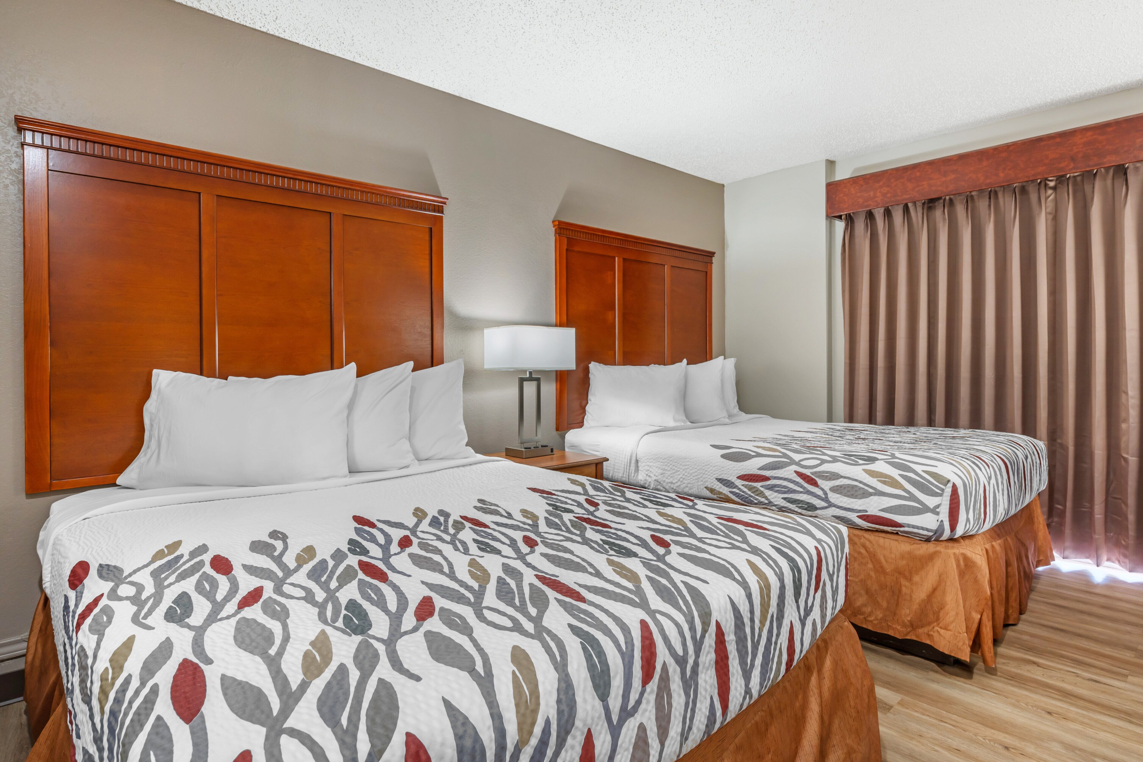 A hotel room with two beds, wooden headboards, a nightstand with a lamp, and a window with curtains.