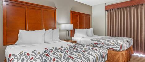 A hotel room with two beds, wooden headboards, a nightstand with a lamp, and a window with curtains.