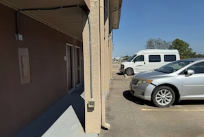 Uma área de estacionamento com um edifício à esquerda e uma carrinha branca e um carro prateado estacionados à direita.