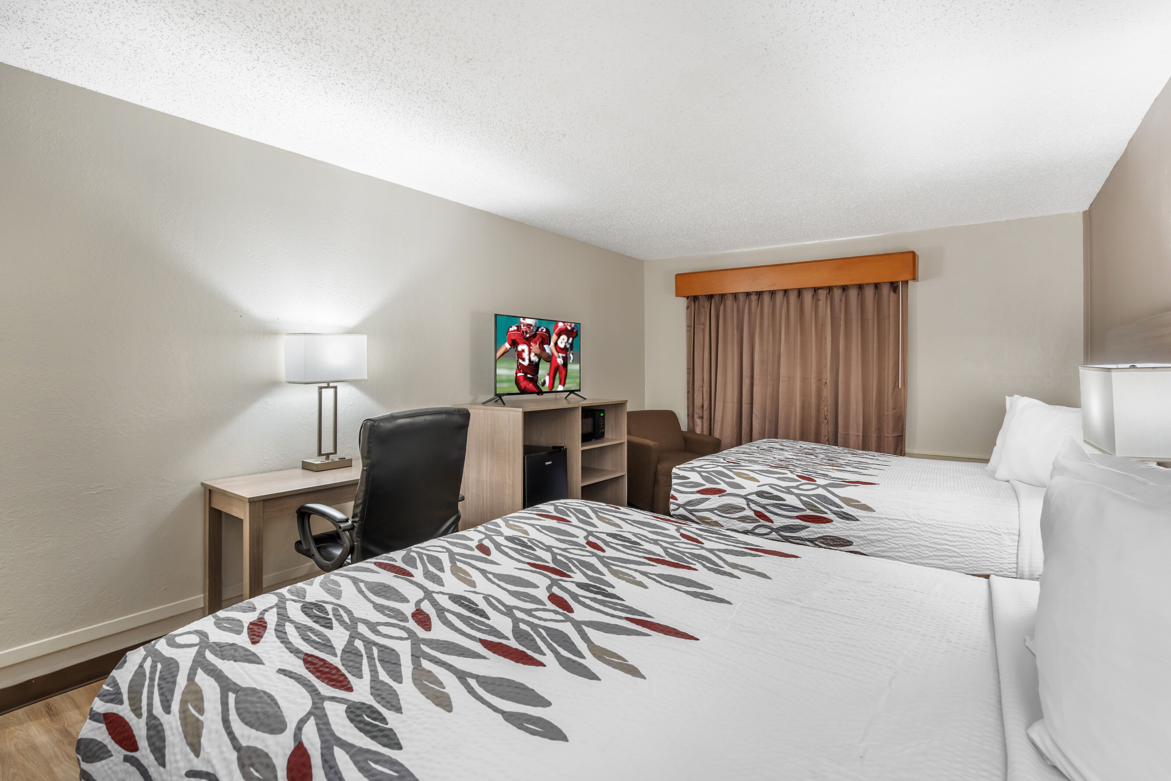 A hotel room with two beds, a desk with a chair, a TV on a stand, and a window with curtains.