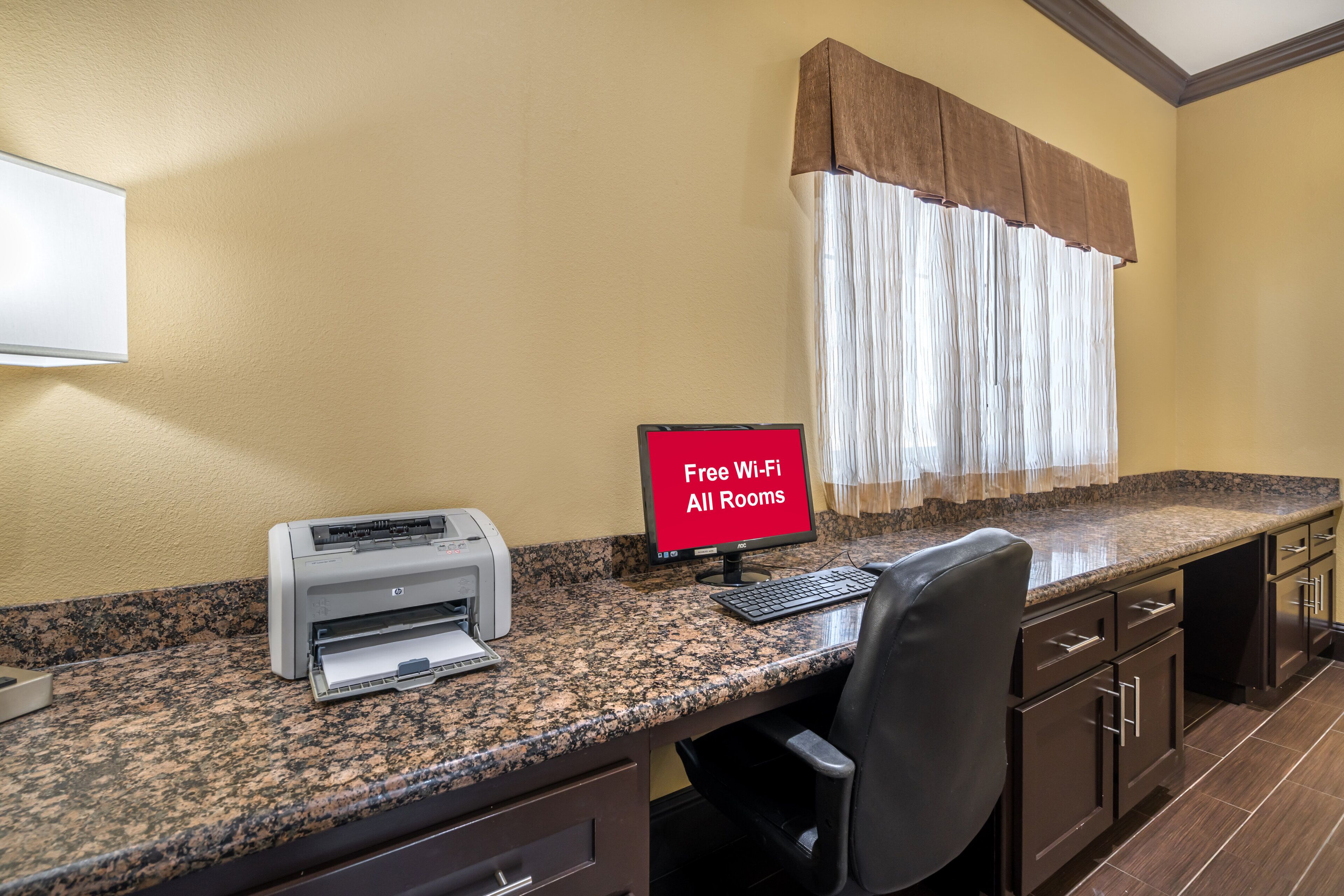 A workspace with a printer, a computer monitor displaying 'Free Wi-Fi All Rooms,' and a window with curtains.