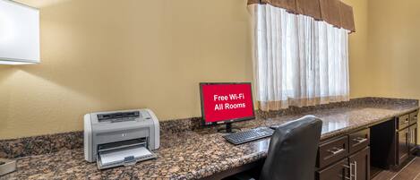 A workspace with a printer, a computer monitor displaying 'Free Wi-Fi All Rooms,' and a window with curtains.