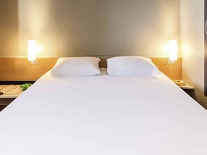 Una habitación de hotel con una cama grande, dos almohadas, una mesita de noche con lámpara y un aplique de pared.