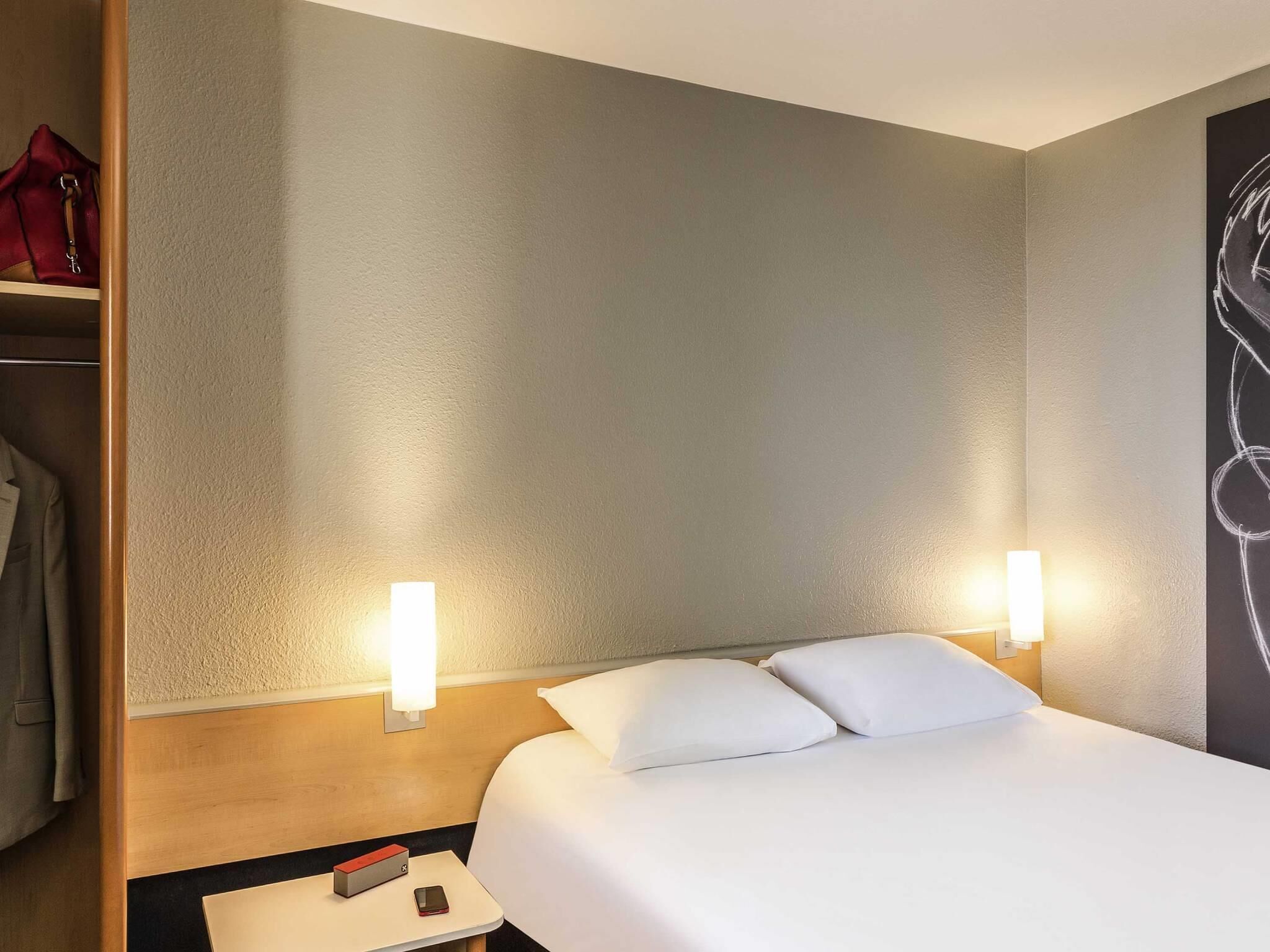 A hotel room with a bed, two bedside lamps, a nightstand with a phone and a red object, and a closet with a red bag.