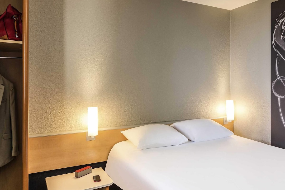 A hotel room with a bed, two bedside lamps, a nightstand with a phone and a red object, and a closet with a red bag.