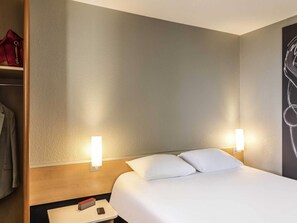 Une chambre d’hôtel avec un lit, deux lampes de chevet, une table de chevet avec un téléphone et un objet rouge, et une armoire contenant un sac rouge.