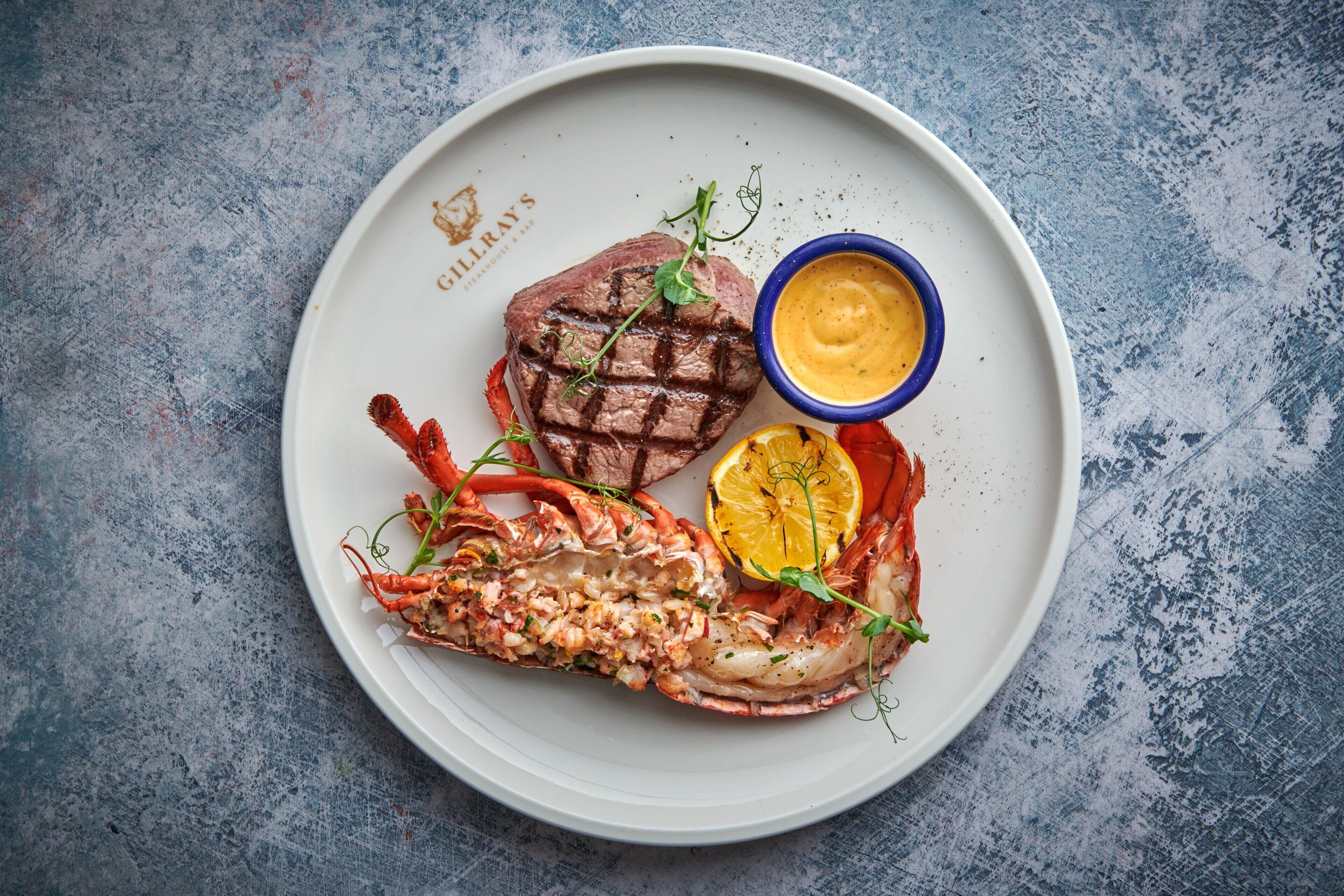 A plate with a grilled steak, lobster tail, and lemon slice, accompanied by a sauce.
