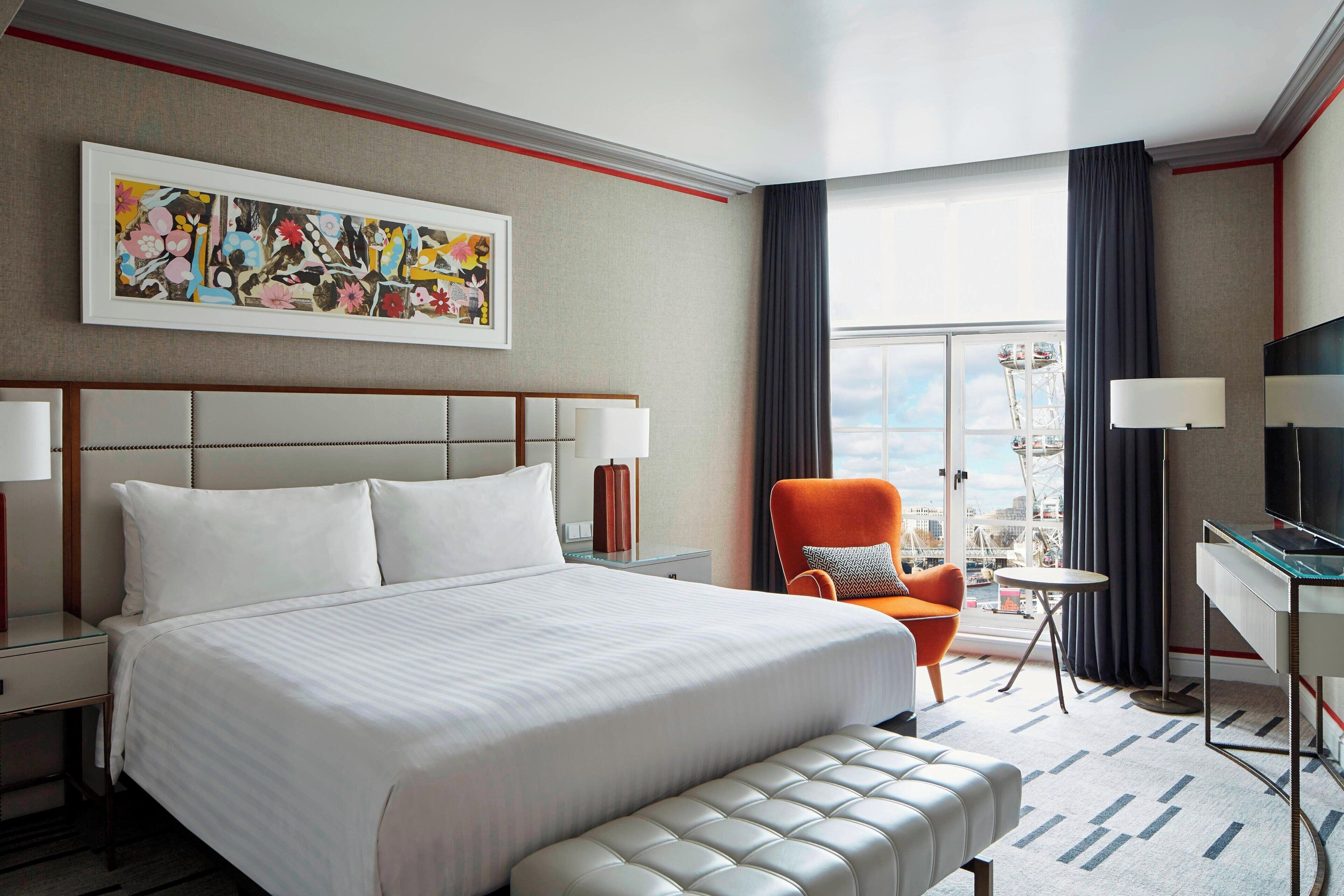 A hotel room with a large bed, a chair, a desk, and a window with a view of a cityscape.
