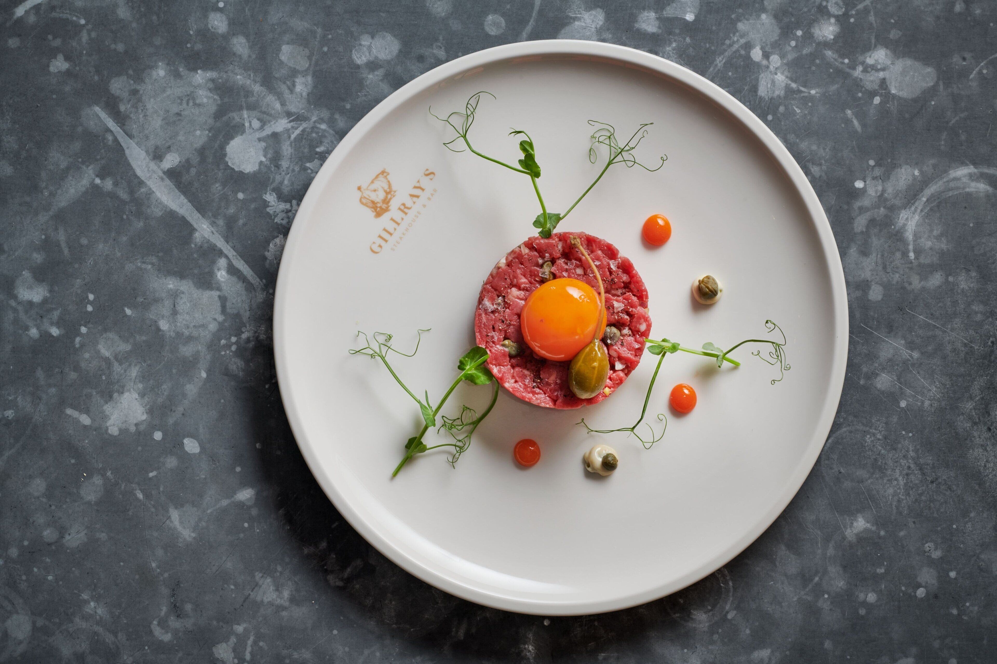 A white plate with a raw meat dish, garnished with cherry tomatoes, olives, and green leaves on a textured gray surface.