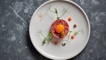 A white plate with a raw meat dish, garnished with cherry tomatoes, olives, and green leaves on a textured gray surface.