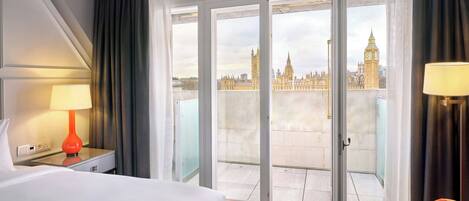 A hotel room with a bed, a nightstand with a lamp, a chair, and a balcony with a view of a historic building.