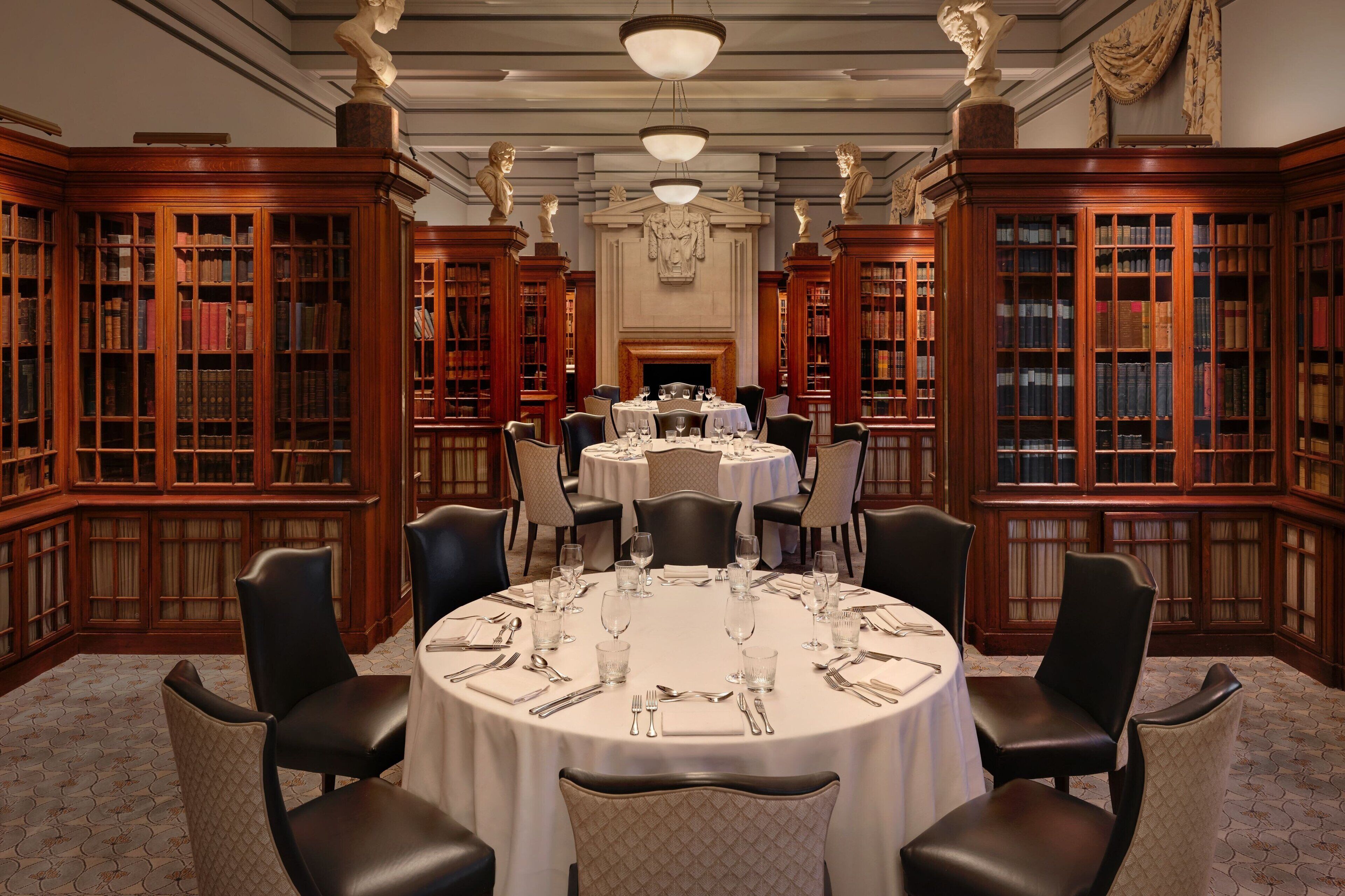 A formal dining room with a round table set for six, surrounded by bookshelves and classical sculptures.