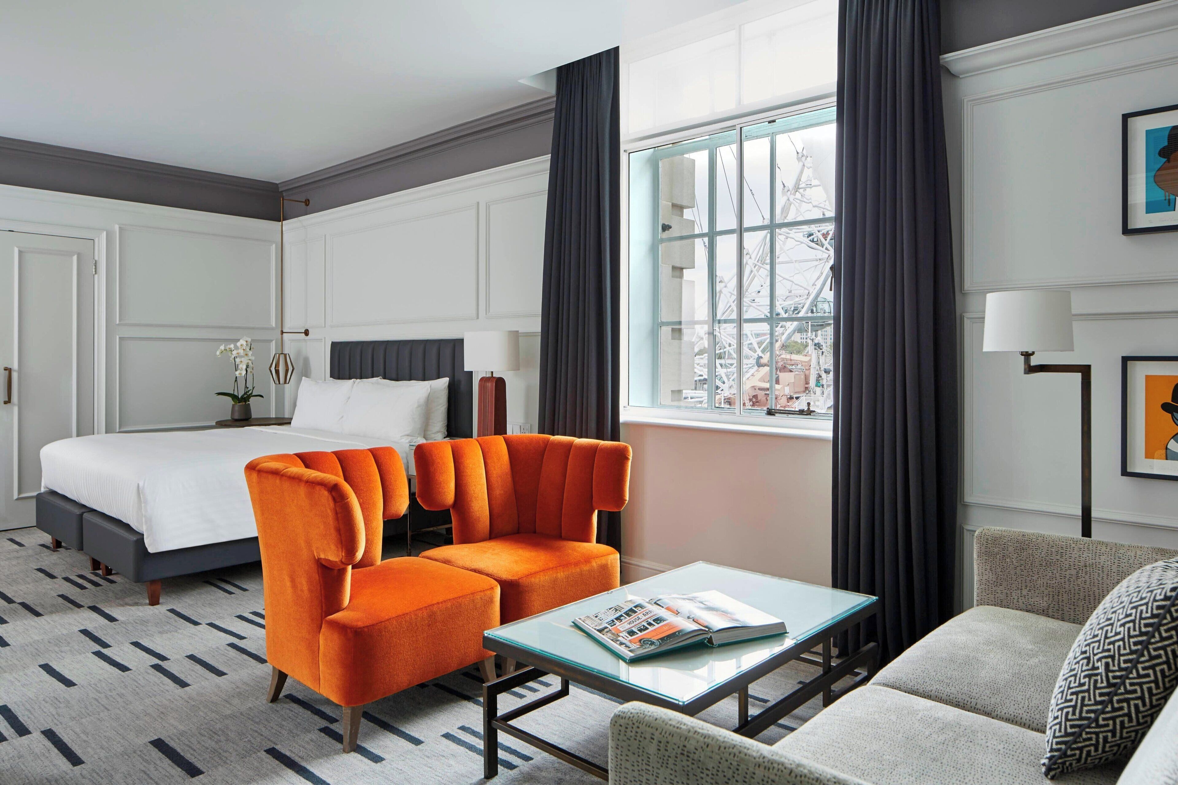 A hotel room with a bed, two orange armchairs, a sofa, a glass coffee table, and a large window with curtains.