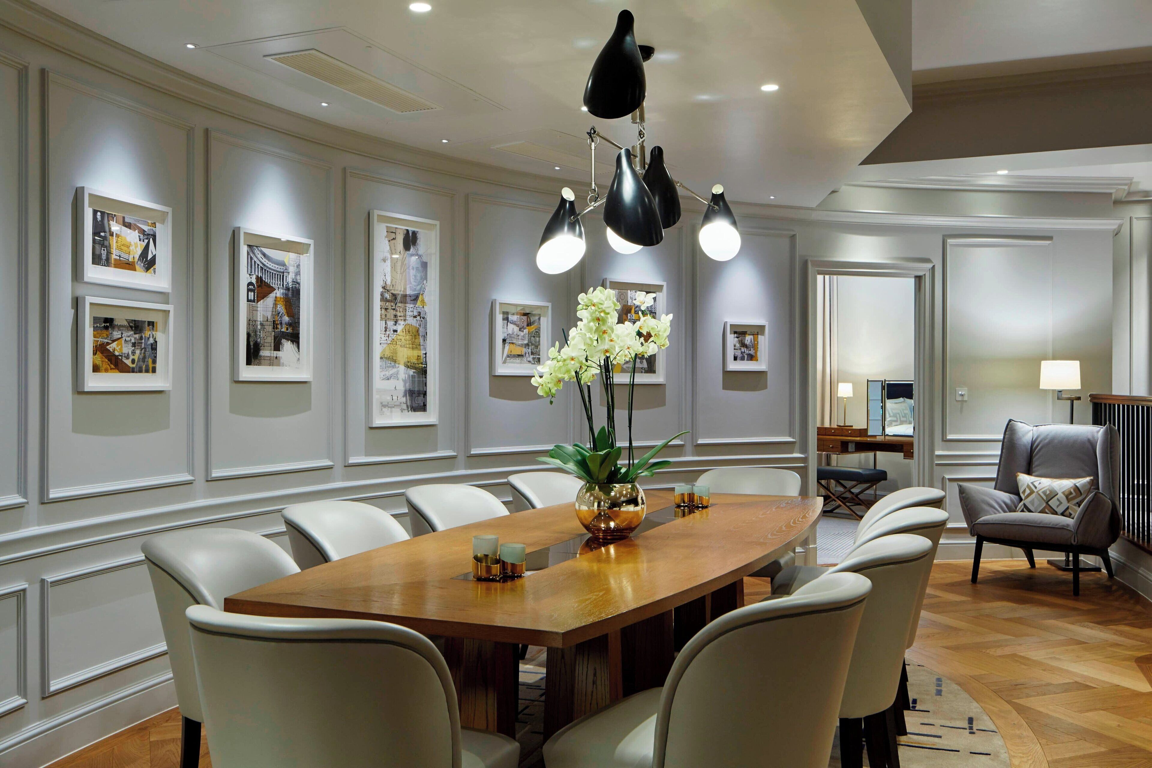 A spacious dining area with a long wooden table surrounded by chairs, a chandelier, framed artwork on the walls, and a vase with flowers on the table.