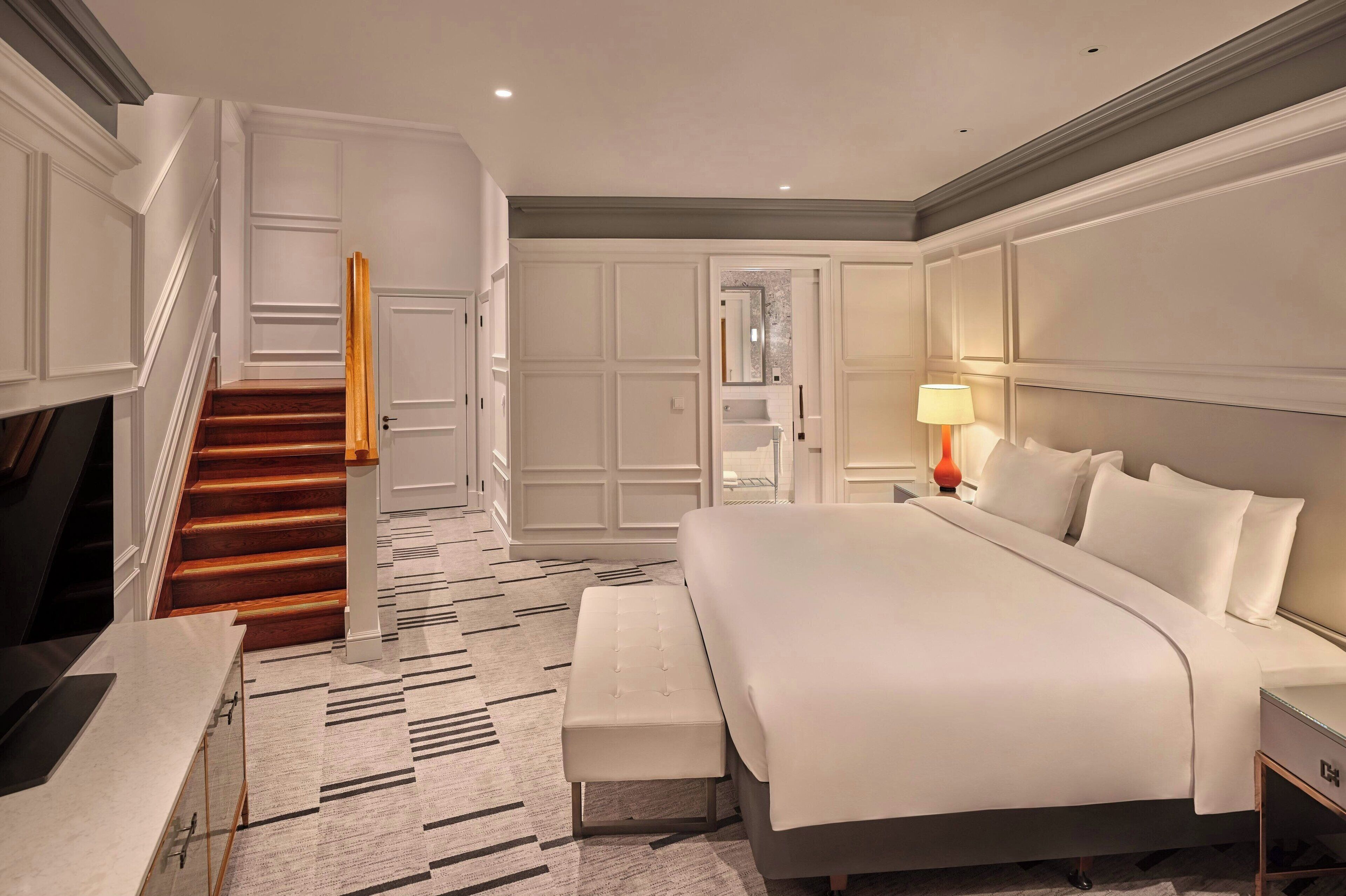 A hotel room with a large bed, a bench, a TV, and a staircase.