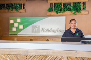 A reception desk with a Holiday Inn sign, wooden paneling, and green foliage decorations. - Holiday Inn Canyon De Chelly by IHG (Chinle)