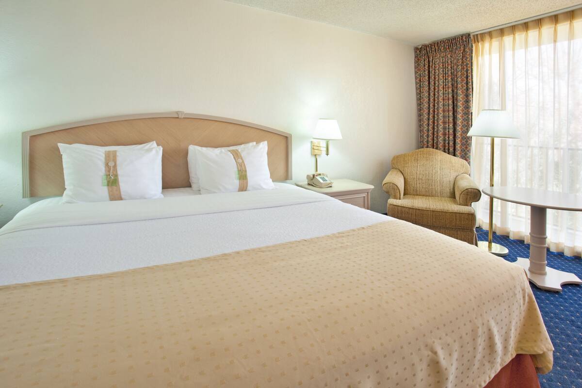 A hotel room with a large bed, a chair, a small table, and a lamp.
