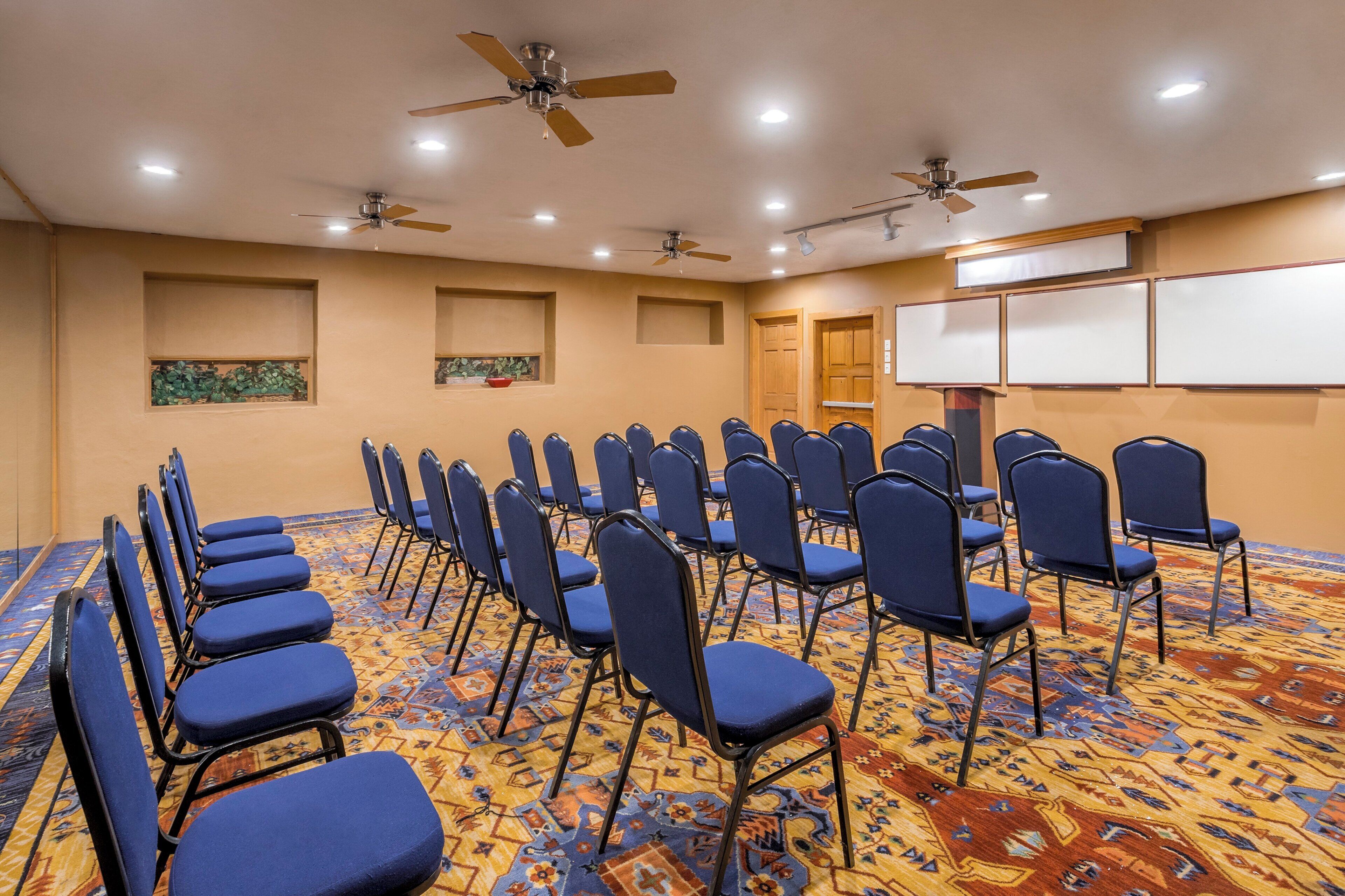 A conference room with rows of blue chairs, ceiling fans, recessed lighting, and a patterned carpet.