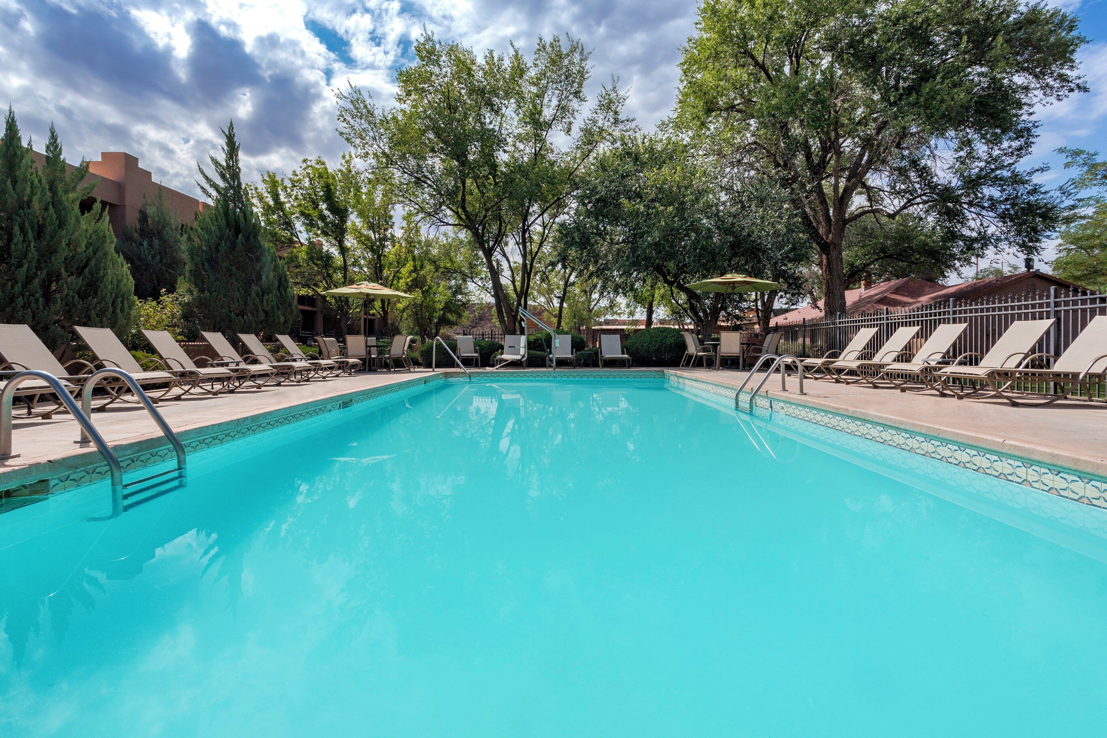 A swimming pool with lounge chairs and umbrellas, surrounded by trees and buildings.