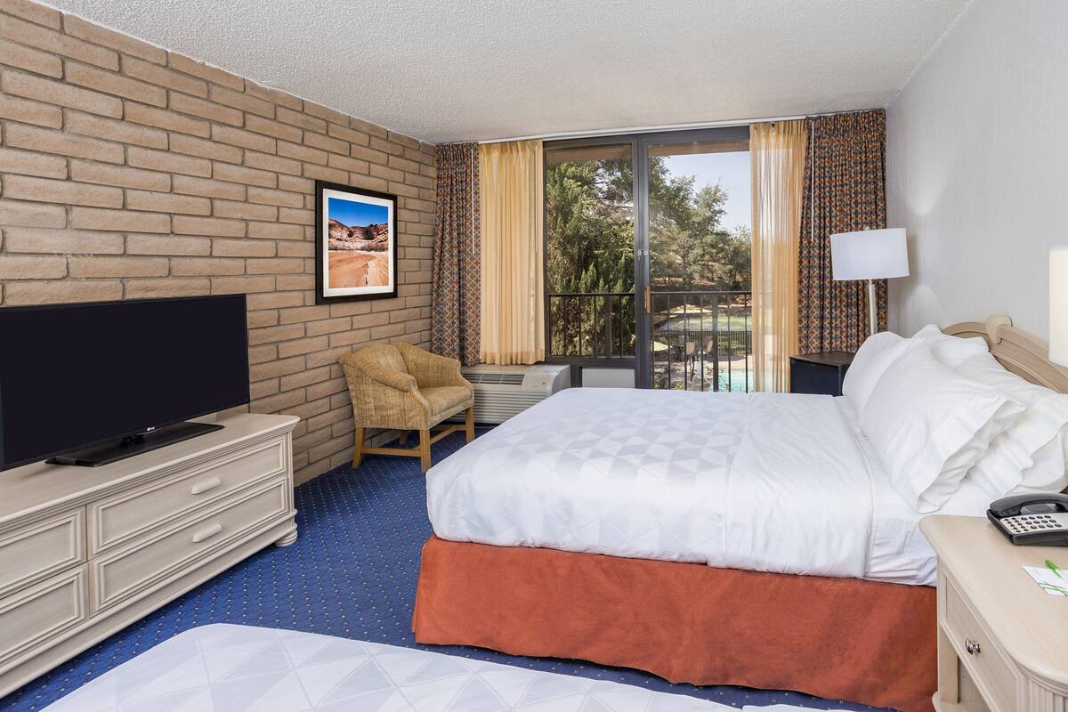 A hotel room with a brick wall, a flat-screen TV, a bed with white bedding, a chair, and a balcony with curtains.