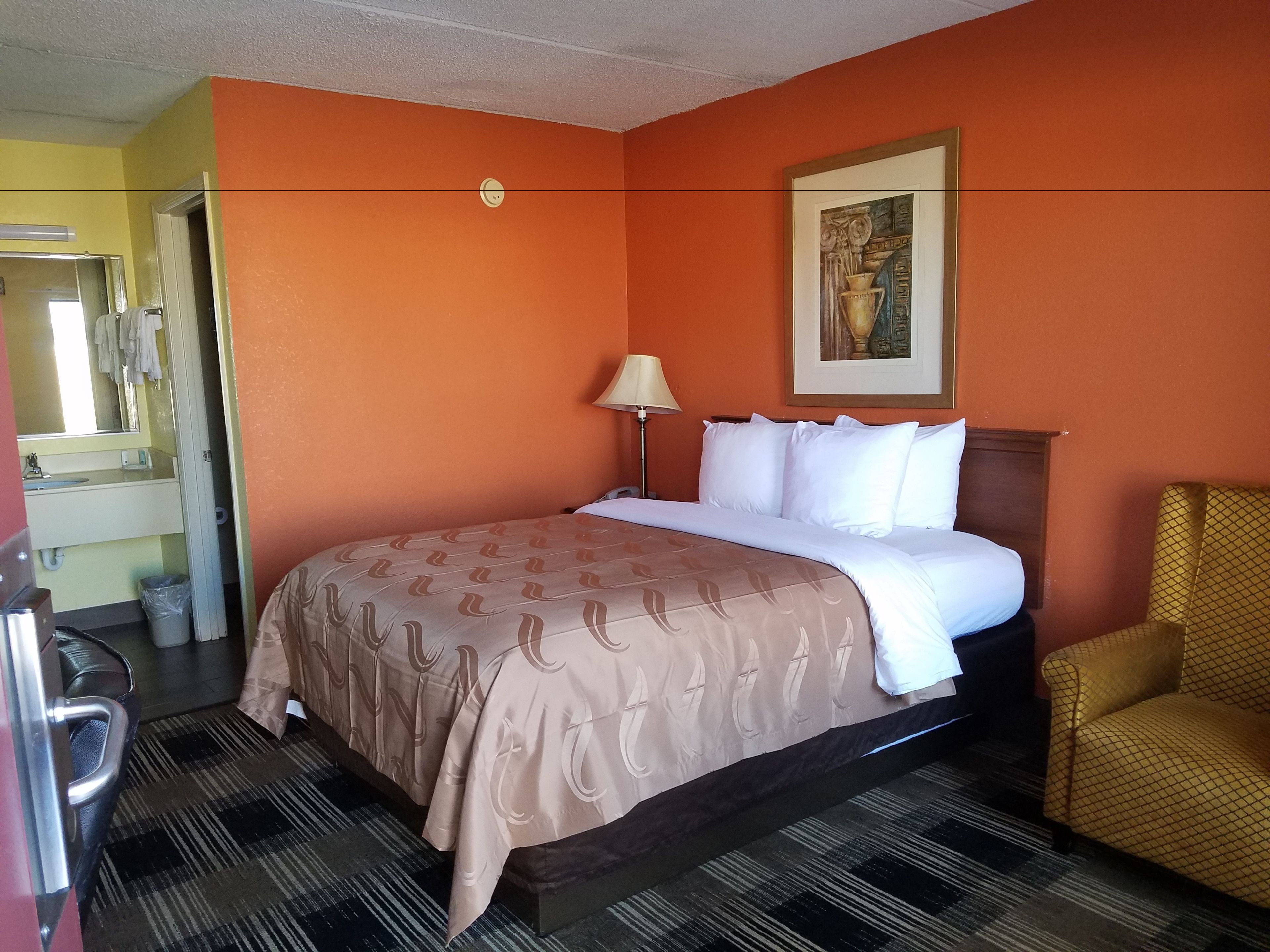 Quarto de hotel com parede laranja, cama com roupa de cama branca, luminária, cadeira e um quadro na parede.