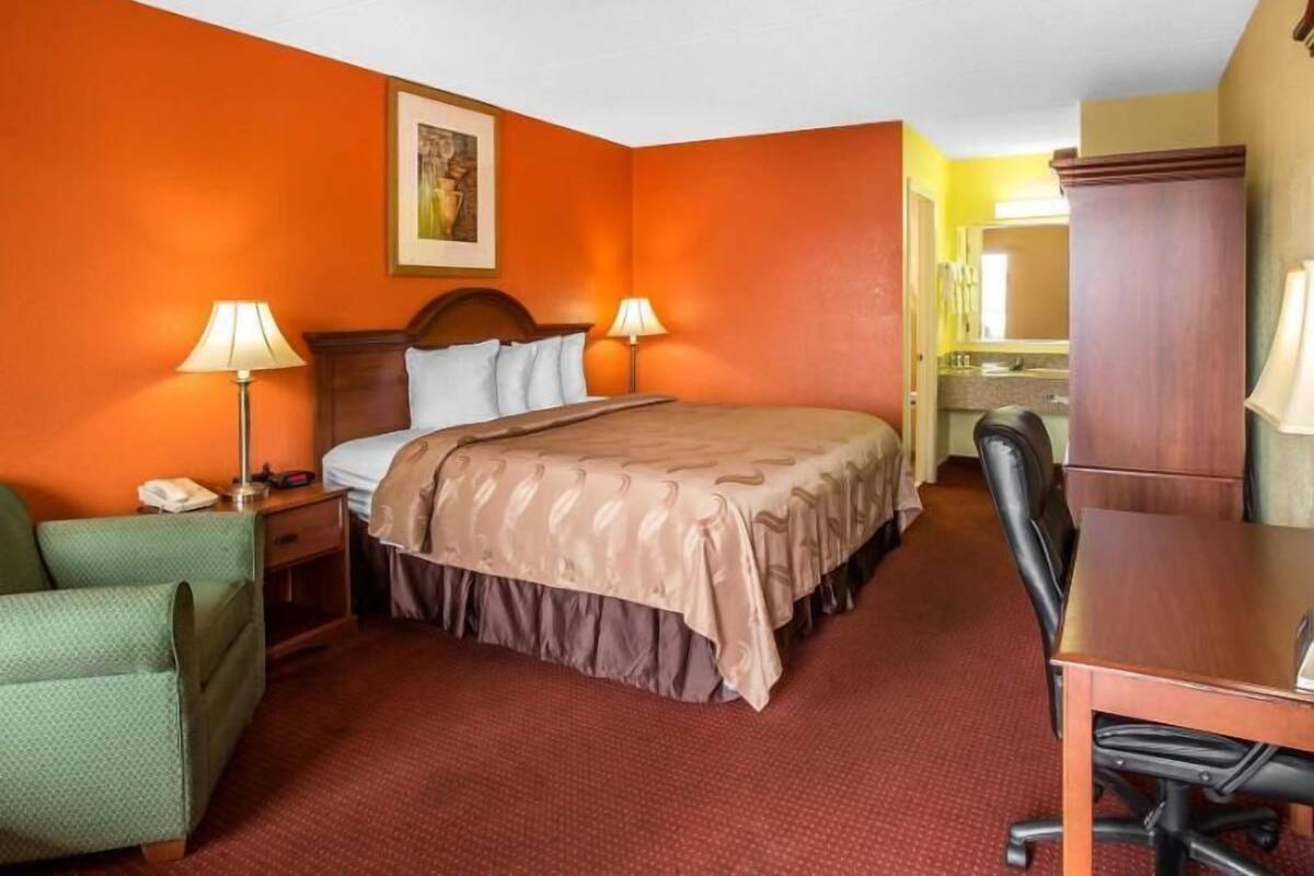 Habitación de hotel con una cama, dos lámparas de noche, un sillón verde, un escritorio de madera y un espejo.
