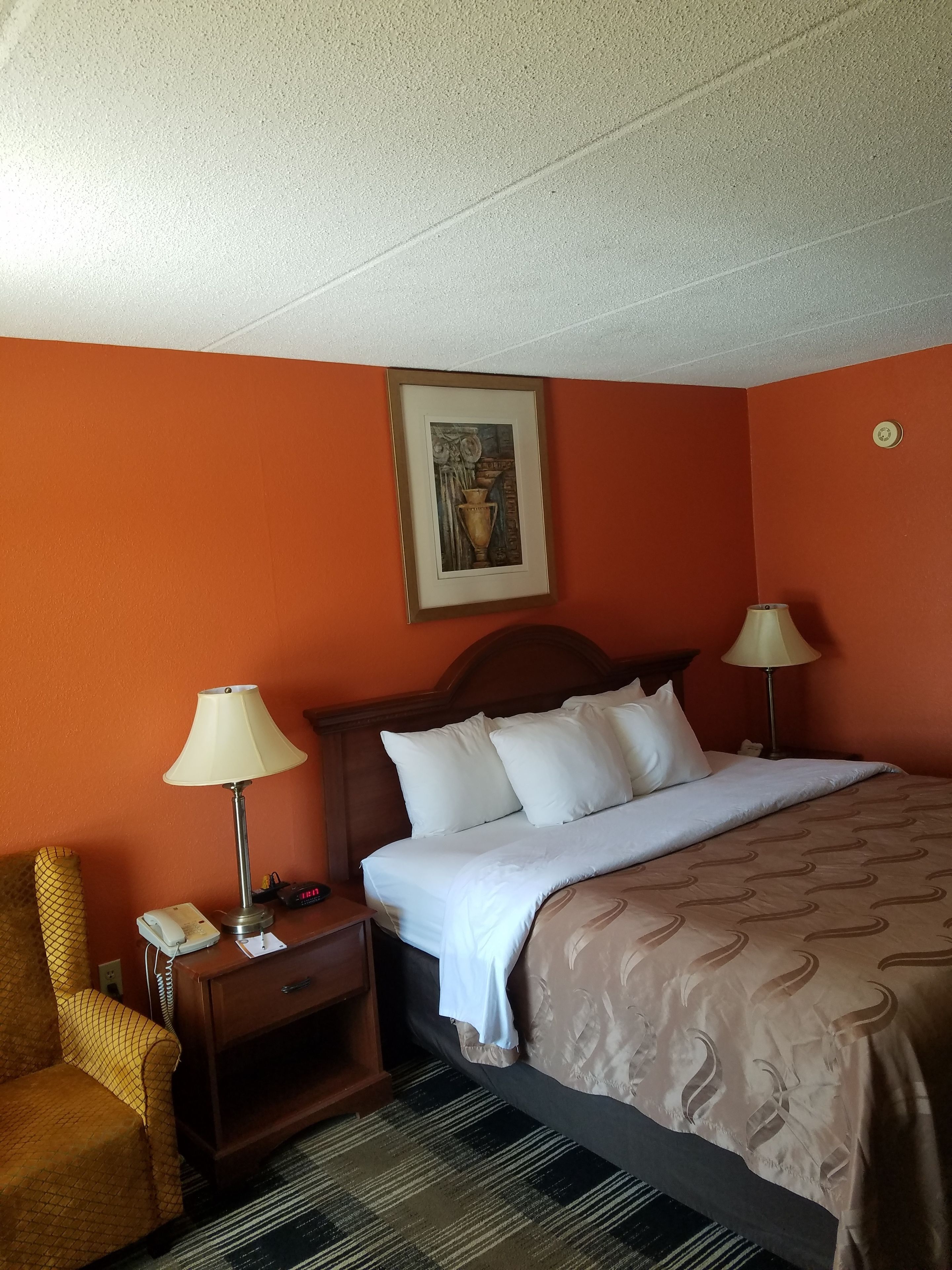 Habitación de hotel con pared naranja, cabecero de madera, cama con ropa blanca, dos lámparas de noche, un teléfono, una mesita y un cuadro enmarcado en la pared.