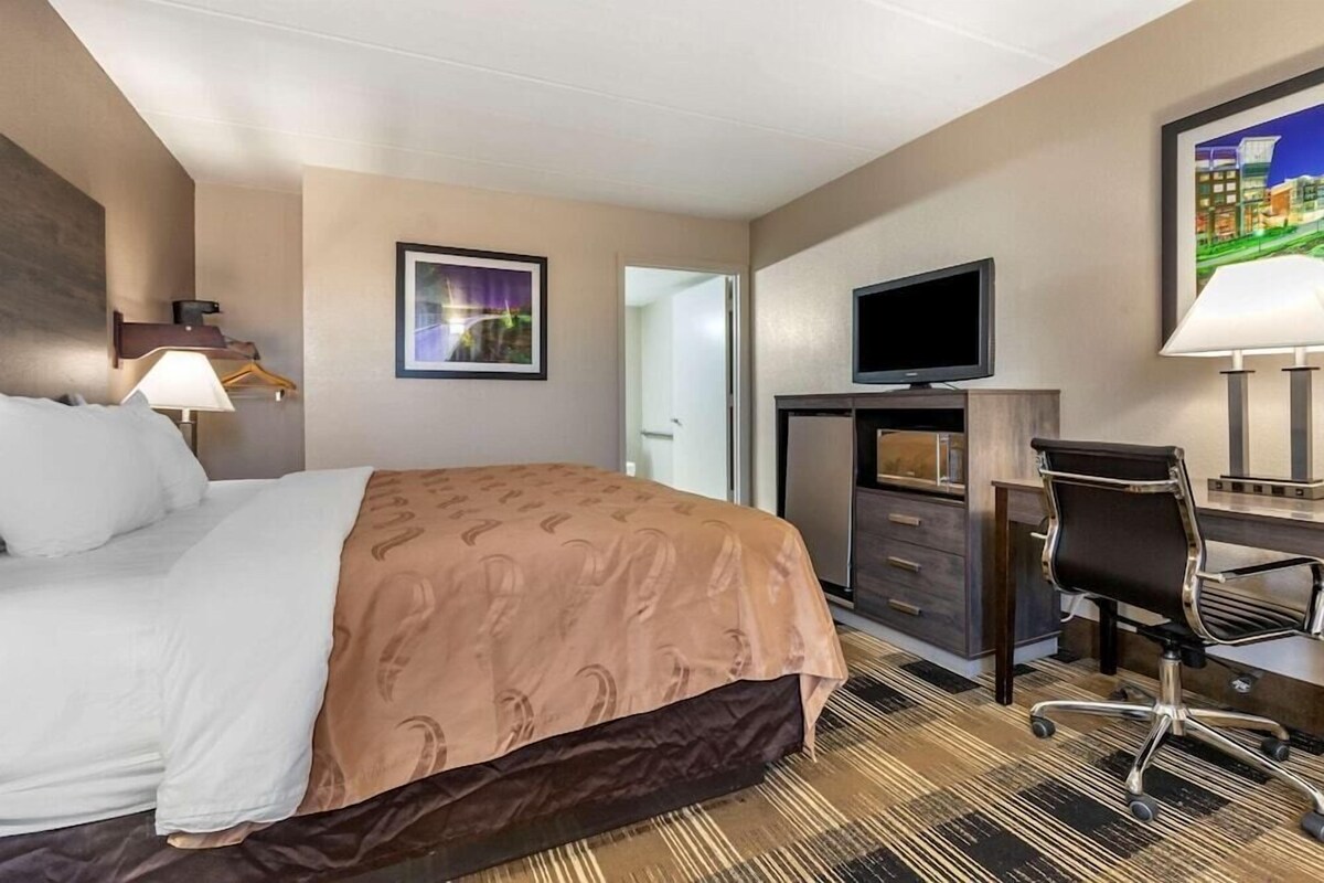 A hotel room with a bed, desk, chair, TV, and a painting on the wall.
