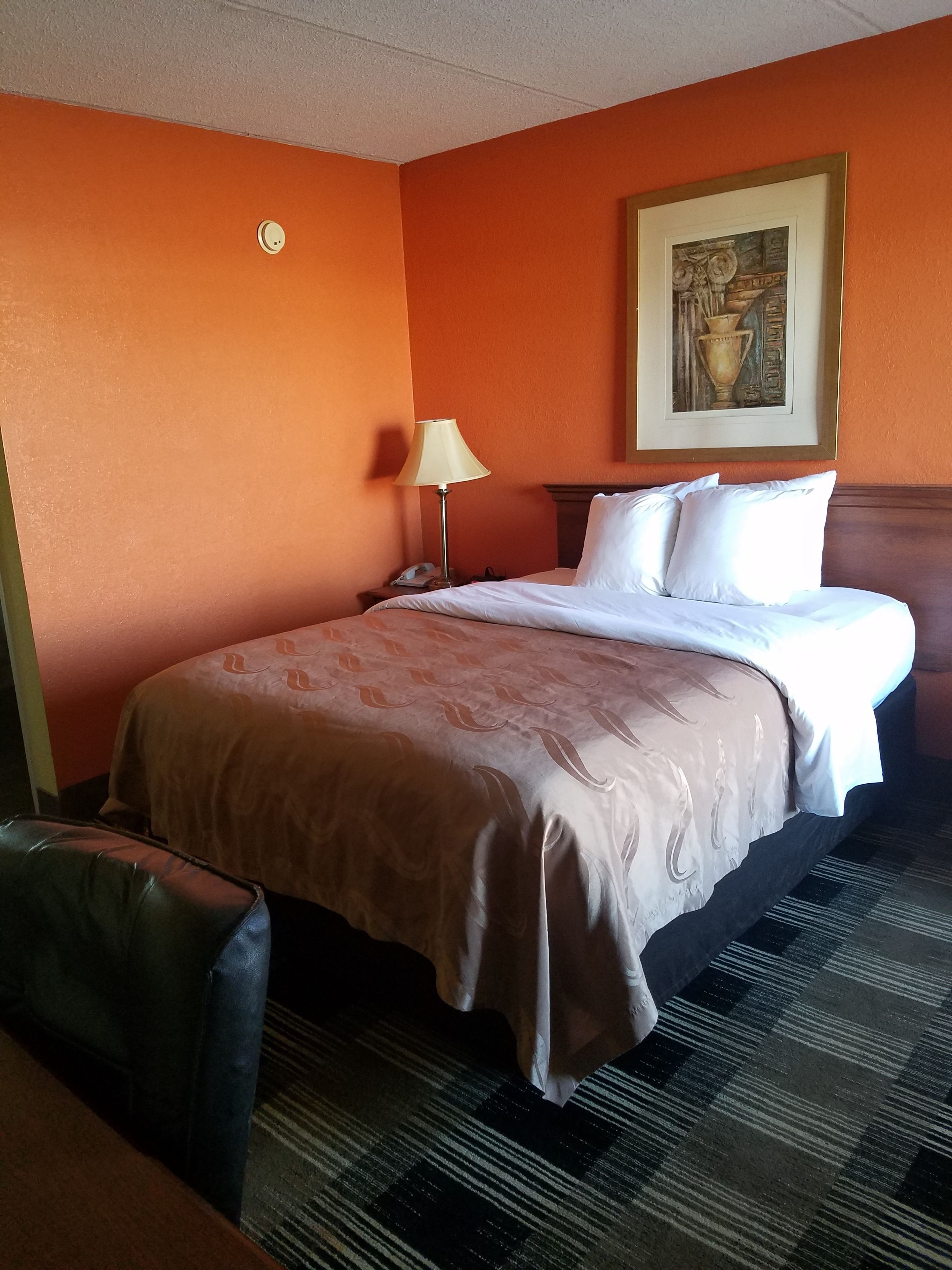 Une chambre d’hôtel avec un mur orange, un lit avec du linge de maison blanc, une lampe et un tableau encadré accroché au mur.