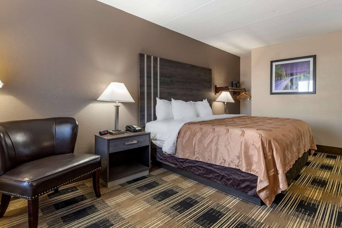 Una habitación de hotel con cama, una silla, una mesita de noche y una lámpara de pared.