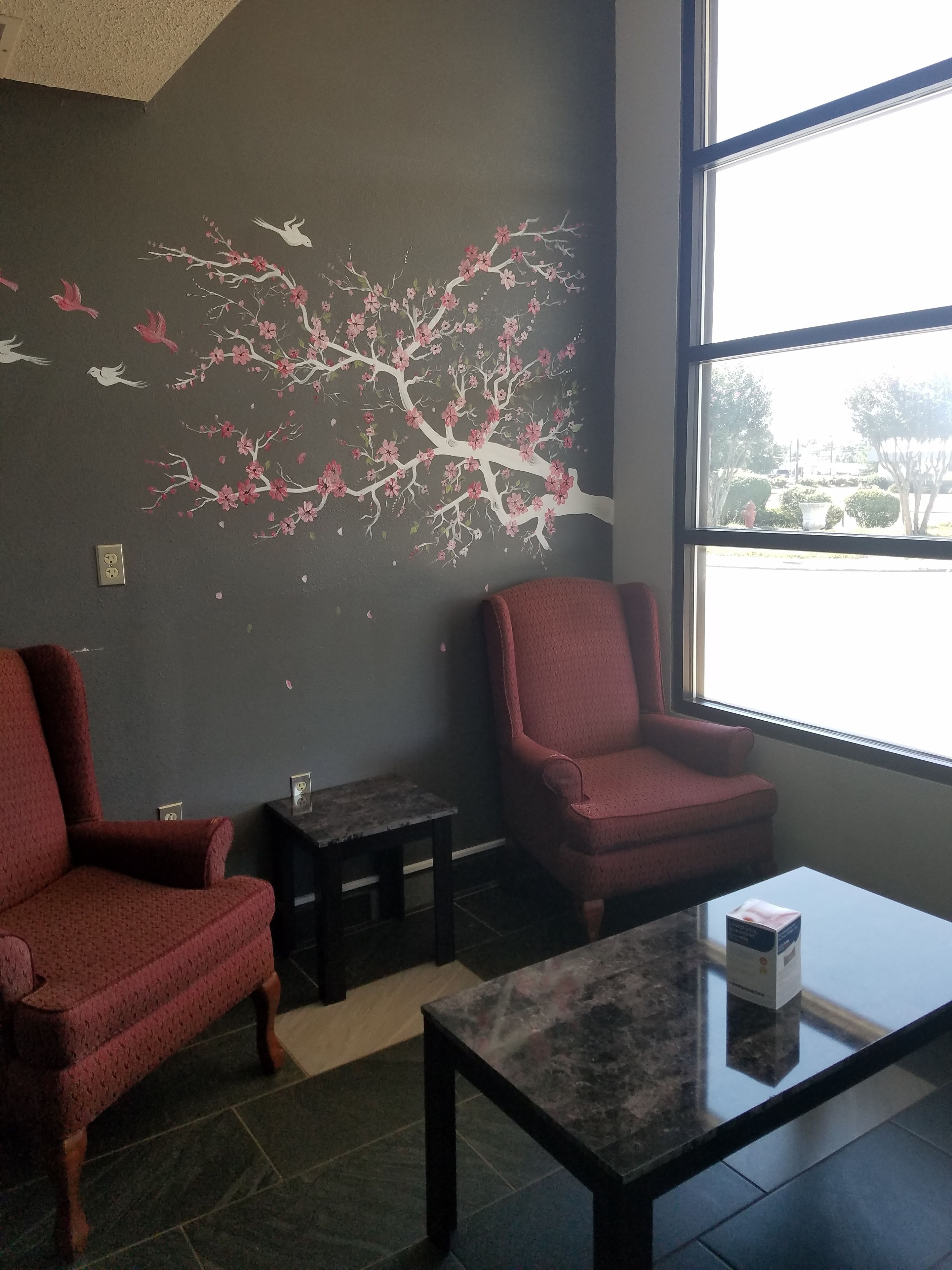 A room with a dark gray wall, a large window, two red armchairs, a small table, and a wall mural of a tree with pink flowers and birds.