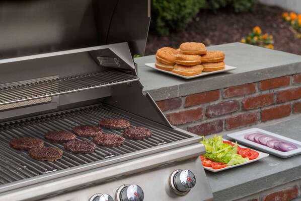 A barbecue grill with burgers cooking, a plate of donuts, and a plate of sliced onions and tomatoes.