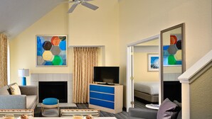 Ein Hotelzimmer mit einer Couch, einem Esstisch mit Frühstück, einem Fernseher, einem Kamin und Kunstwerken an den Wänden.