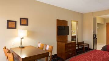 A hotel room with a wooden dining table, chairs, a TV on a wooden cabinet, a bed with a red blanket, and a small table with a chair.